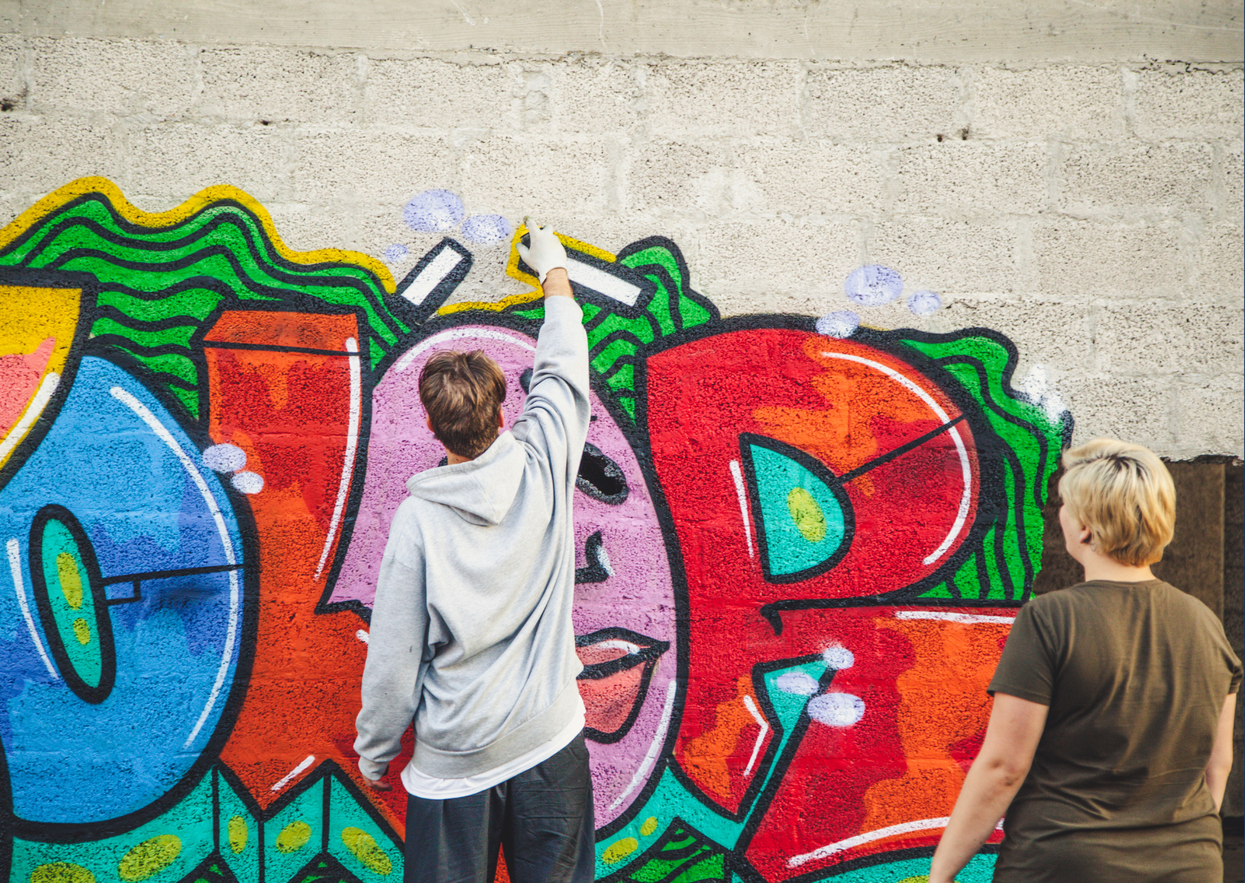 Twee mensen maken graffiti op een muur met kleurrijke kunstwerken en woorden.