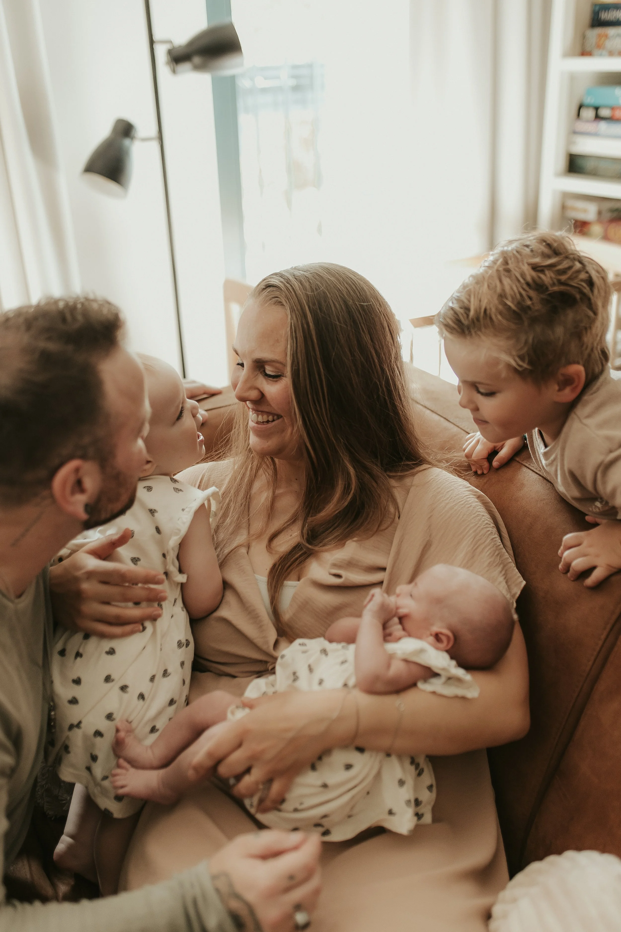 Een vrouw met lang bruin haar zit op een bank, omringd door vier kinderen en twee baby's, allemaal liefdevol samengekomen in een gezellige woonkamer.