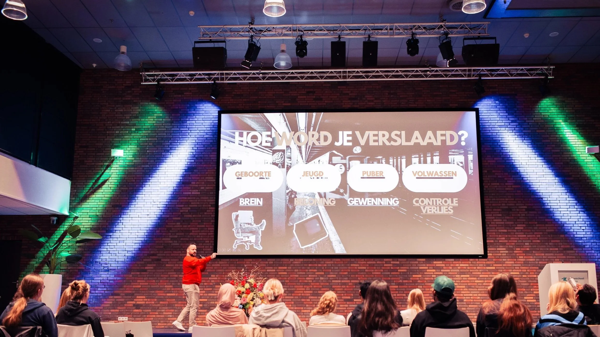 Een spreker geeft een presentatie aan een groep mensen in een zaal met een grote scherm waarop vragen en woorden staan, onder andere over levensfasen en hersenontwikkeling, met kleurrijke verlichting op een bakstenen muur.