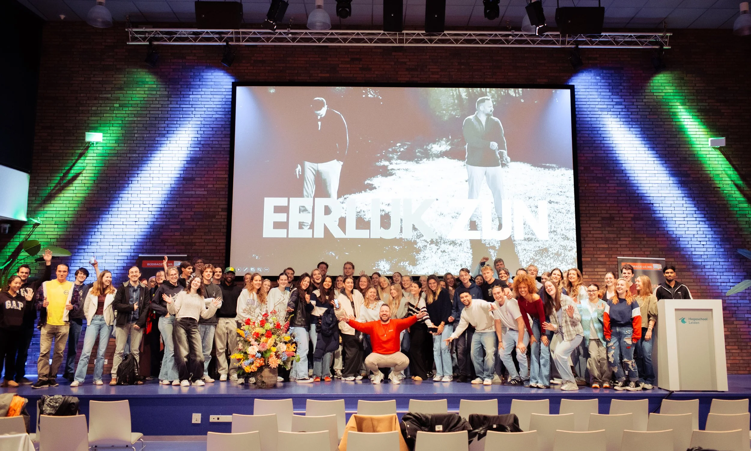 Groep mensen op een podium voor een grote scherm met een zwart-wit afbeelding van twee mannen en de tekst 'EERLIJK ZUIJN'. De groep bestaat uit jonge mensen die zwaaien en glimlachen, met bloemstukken en kleurrijke achtergrondverlichting.