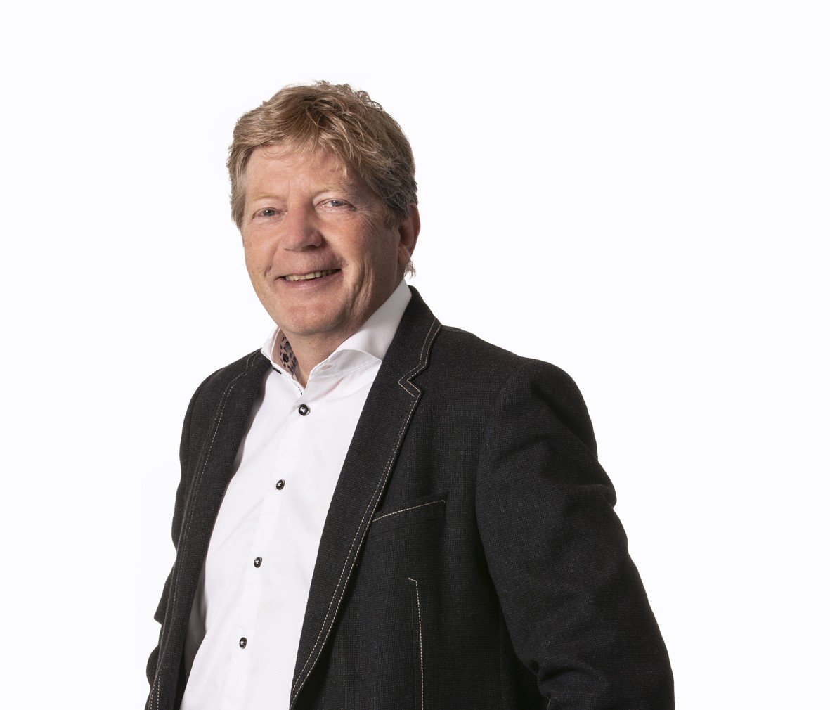 Man met blond haar, glimlachend, draag een witte blouse en een donkere jas, staand tegen een witte achtergrond.