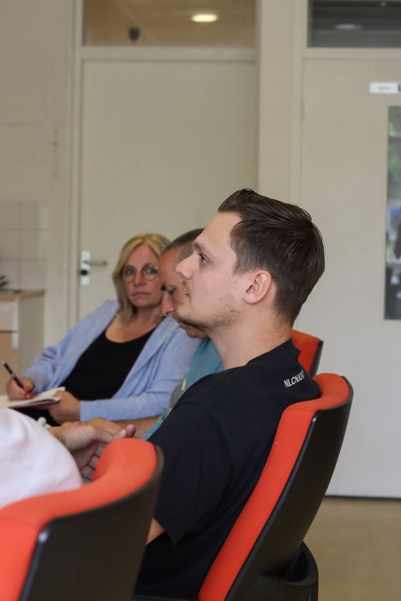 Een jonge man zit aan een tafel in een vergaderruimte, omringd door volwassenen die luisteren en notities maken.