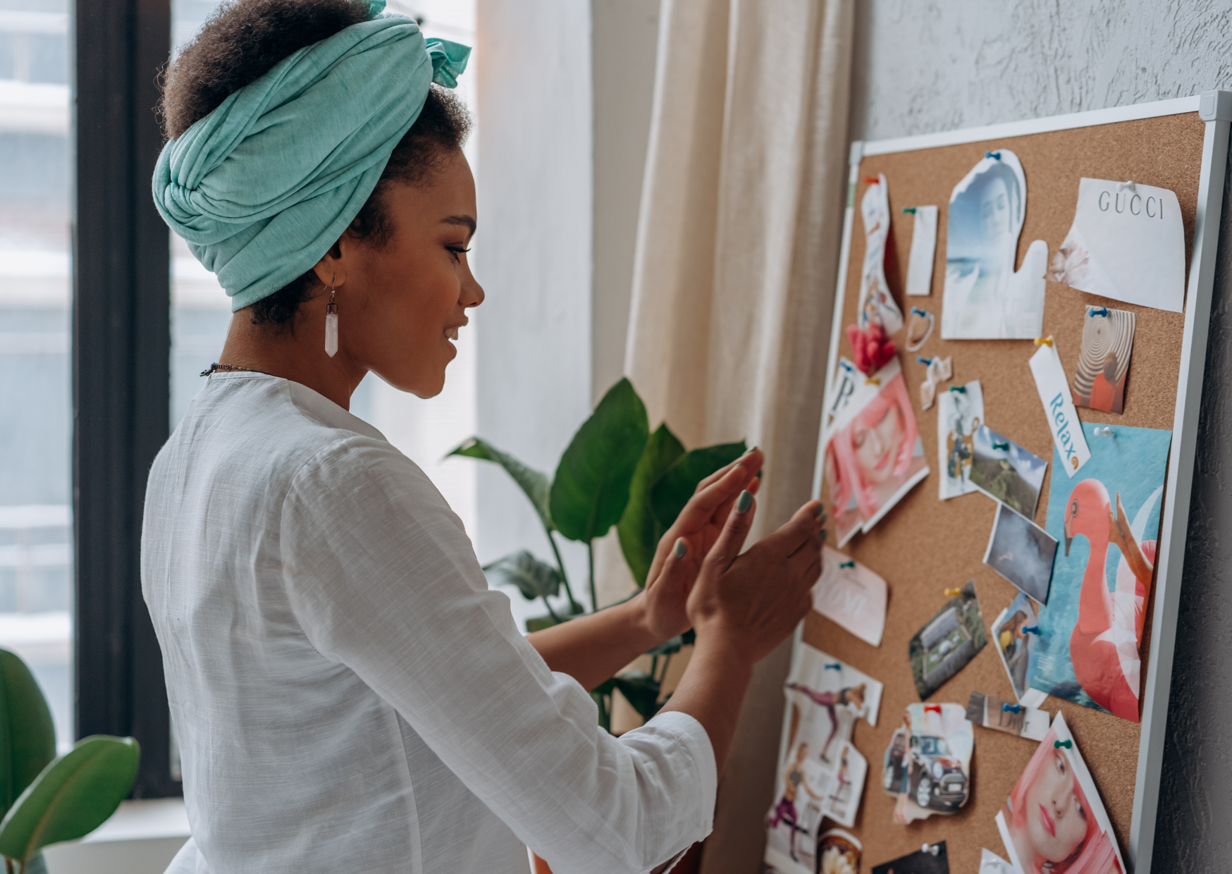 Vrouw met een groene hoofddoek die foto's op een prikbord bevestigt in een kamer met veel natuurlijk licht.