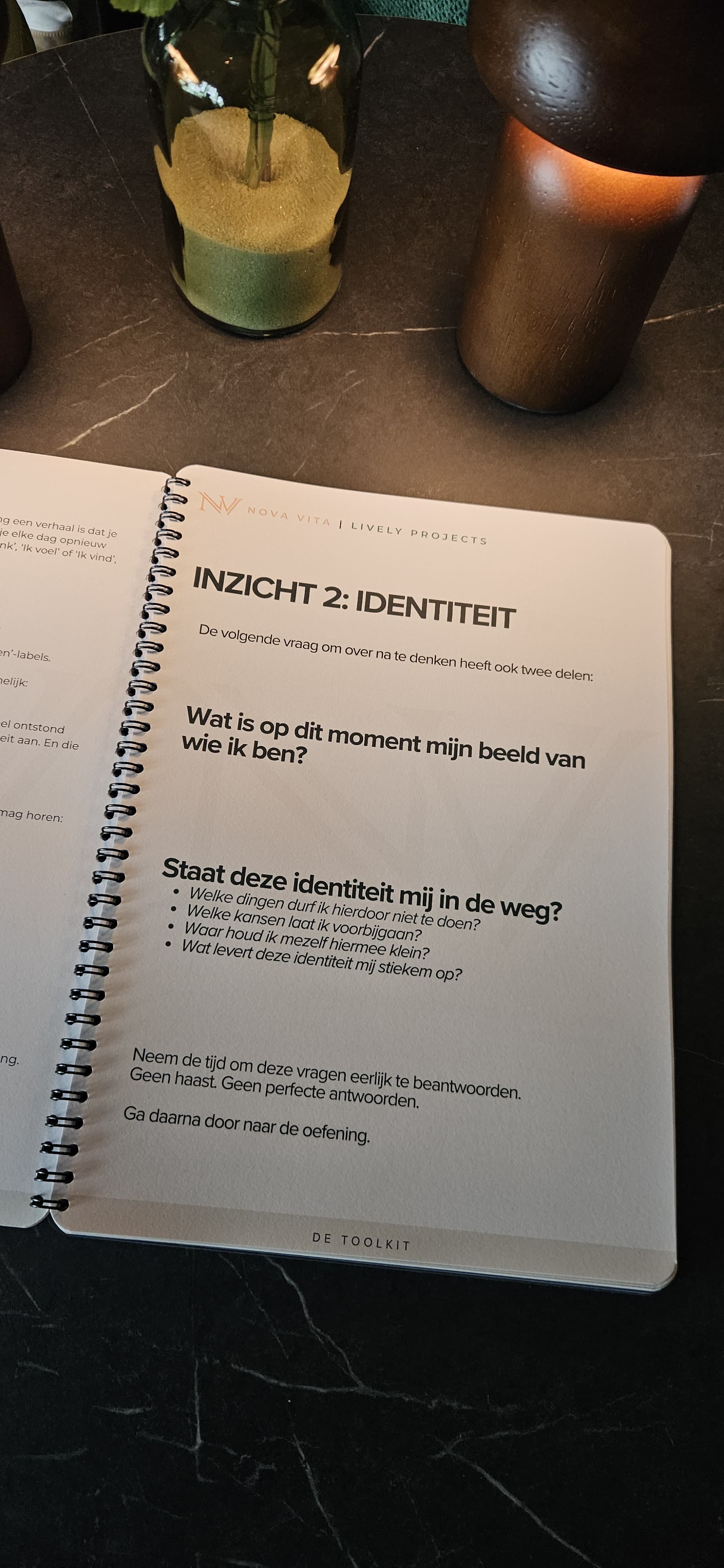 Een open notitieboek op een tafel met een pen, een pot met zand en een lamp ernaast.