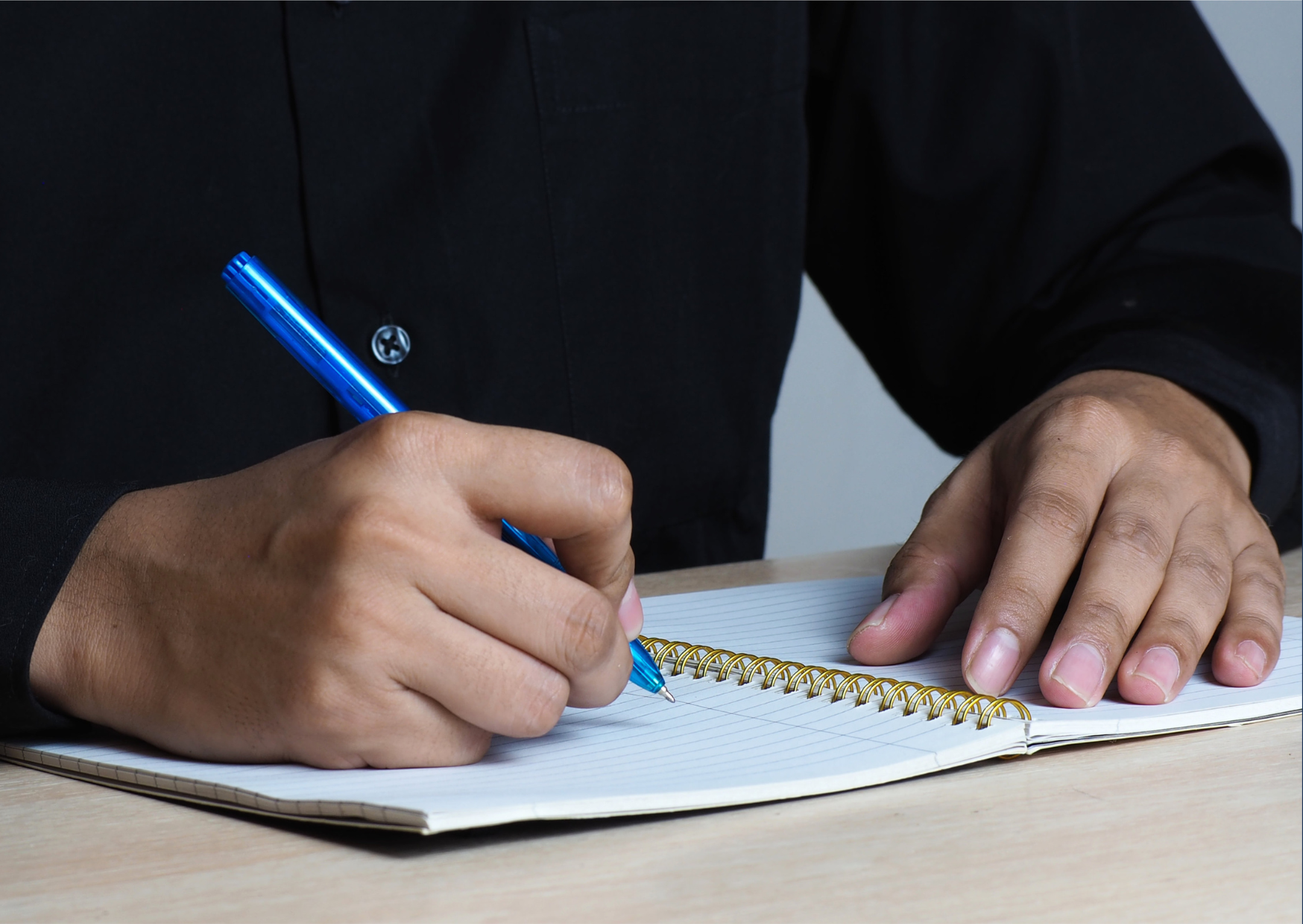 Persoon schrijft met een blauwe pen in een schrift met gewelfde zi-rollen op een tafel.