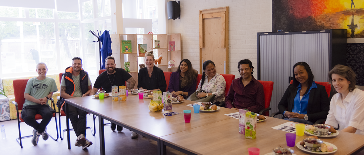Een groep mensen zit aan een lange tafel in een vergaderruimte met grote ramen, er is ontbijt met fruit, drankjes en papieren. Ze lachen en lijken te genieten van een bijeenkomst of teamlunch.