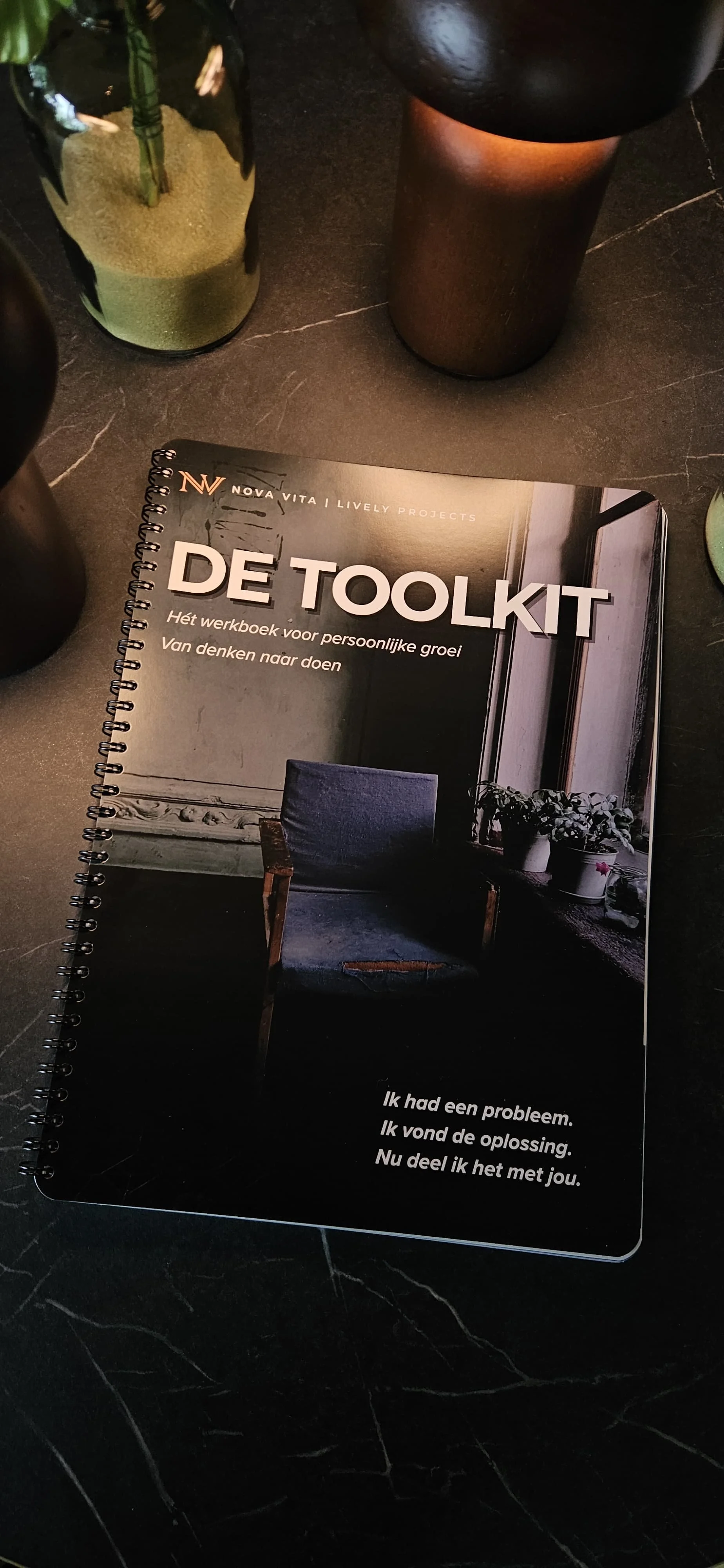 De afbeelding toont een boek genaamd 'De Toolbox' op een donkere marmeren tafel, omringd door een groene vaas en een bruin metallic kan met een zwart bovenstuk.