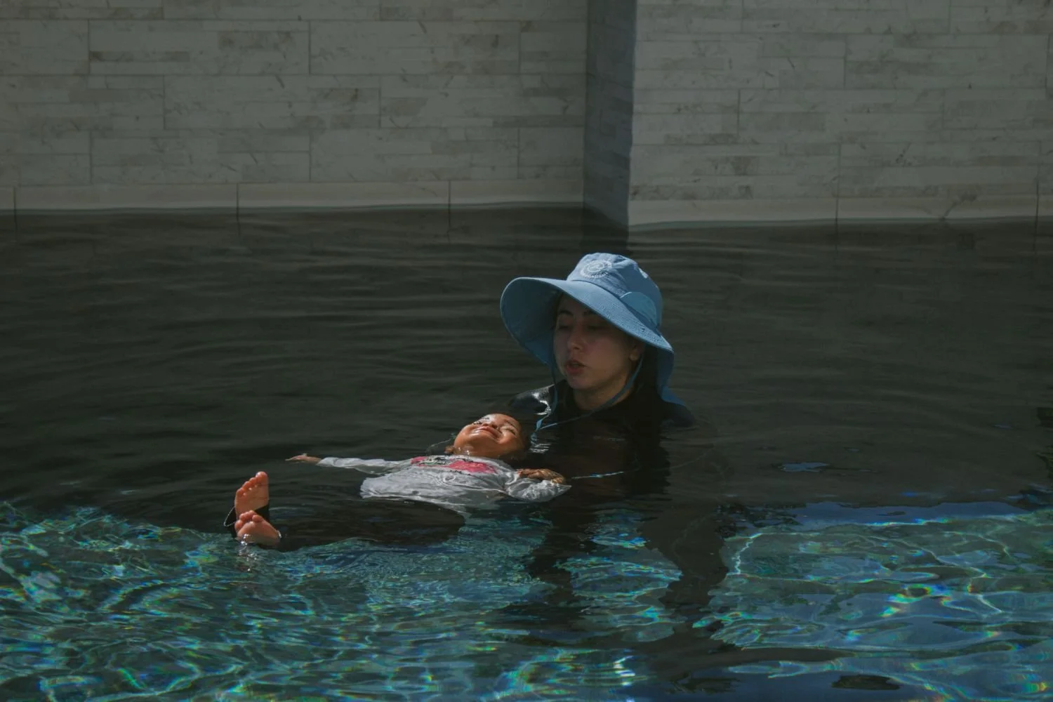 A woman wearing a large blue sun hat is in a swimming pool, holding a young girl who appears to be enjoying the water during her survival swim lesson in California