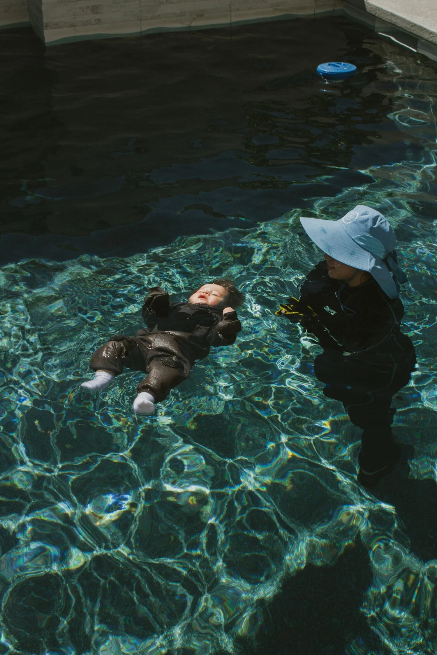 Child floating in swimming pool with a woman, possibly a lifeguard or swimming instructor, standing nearby. The child is wearing a black outfit and white socks, and the woman is wearing a wide-brimmed hat and black clothing.  Survival Swim Lessons