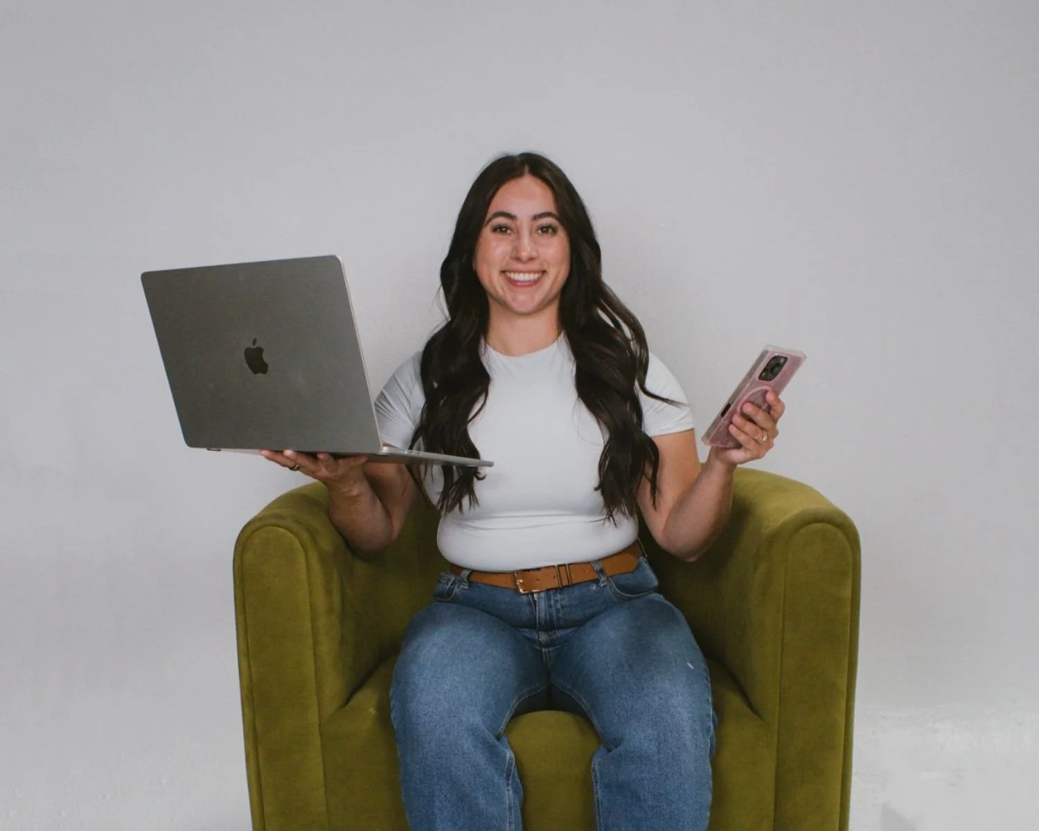 A woman sitting on a green armchair, holding a laptop in her left hand and a smartphone in her right hand, smiling at the camera for Survival Swim Lessons