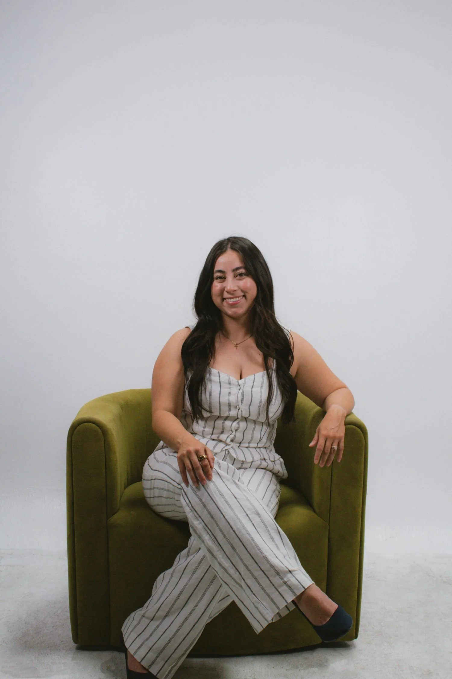 A woman sitting on a green armchair, smiling at the camera, wearing a striped sleeveless jumpsuit with black shoes, in front of a plain white background. Survival Swim Lesson