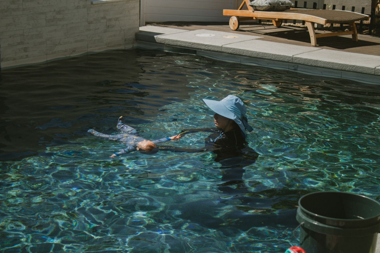 A woman wearing a large sun hat helps a young girl in a swimsuit float in a pool.
