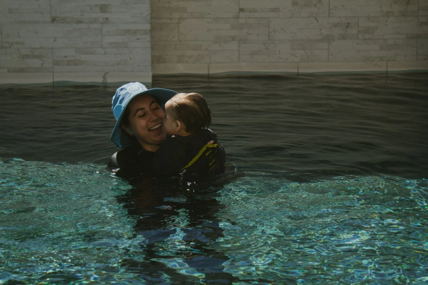 A woman in a blue hat is smiling and holding a young child underwater in a pool with a stone wall background.  Survival Swim Lessons