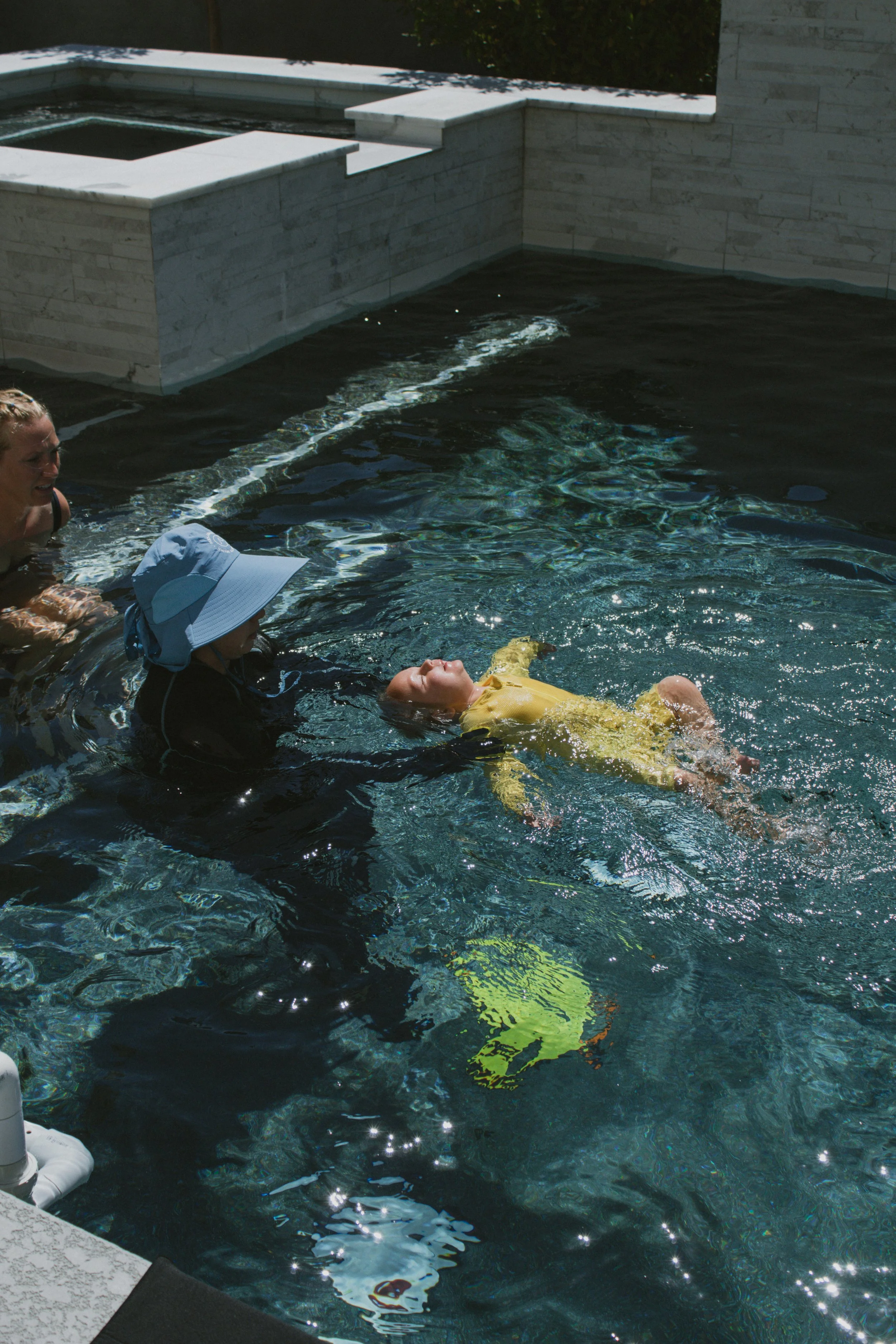 A young child in a yellow outfit is floating on their back in a swimming pool with three adults nearby, helping or supervising. The pool has a modern design with white stone walls.  Survival Swim Lessons
