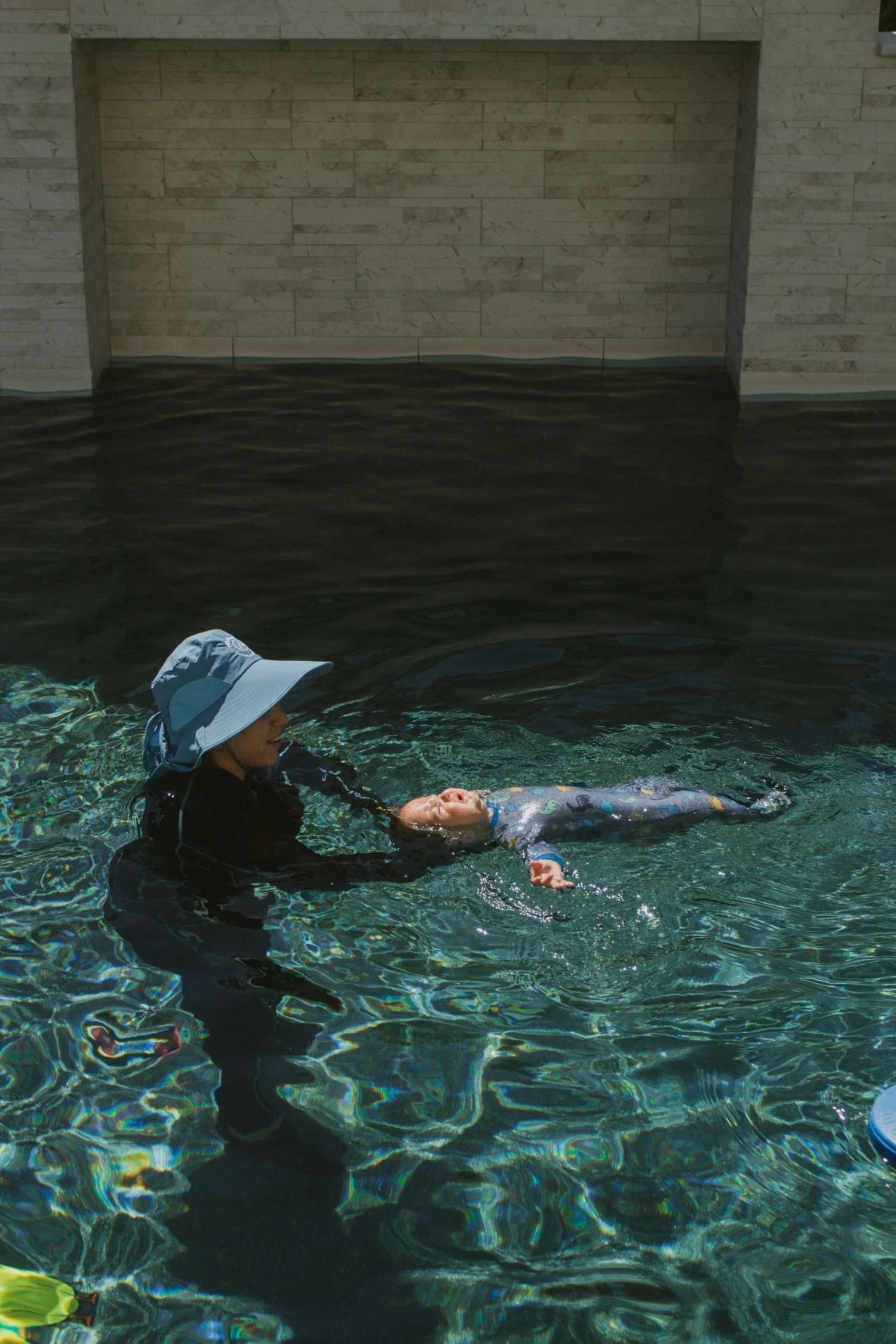 A woman in a wide-brimmed hat and black wetsuit assisting a young child in a patterned wetsuit during a survival swim lesson in a pool.