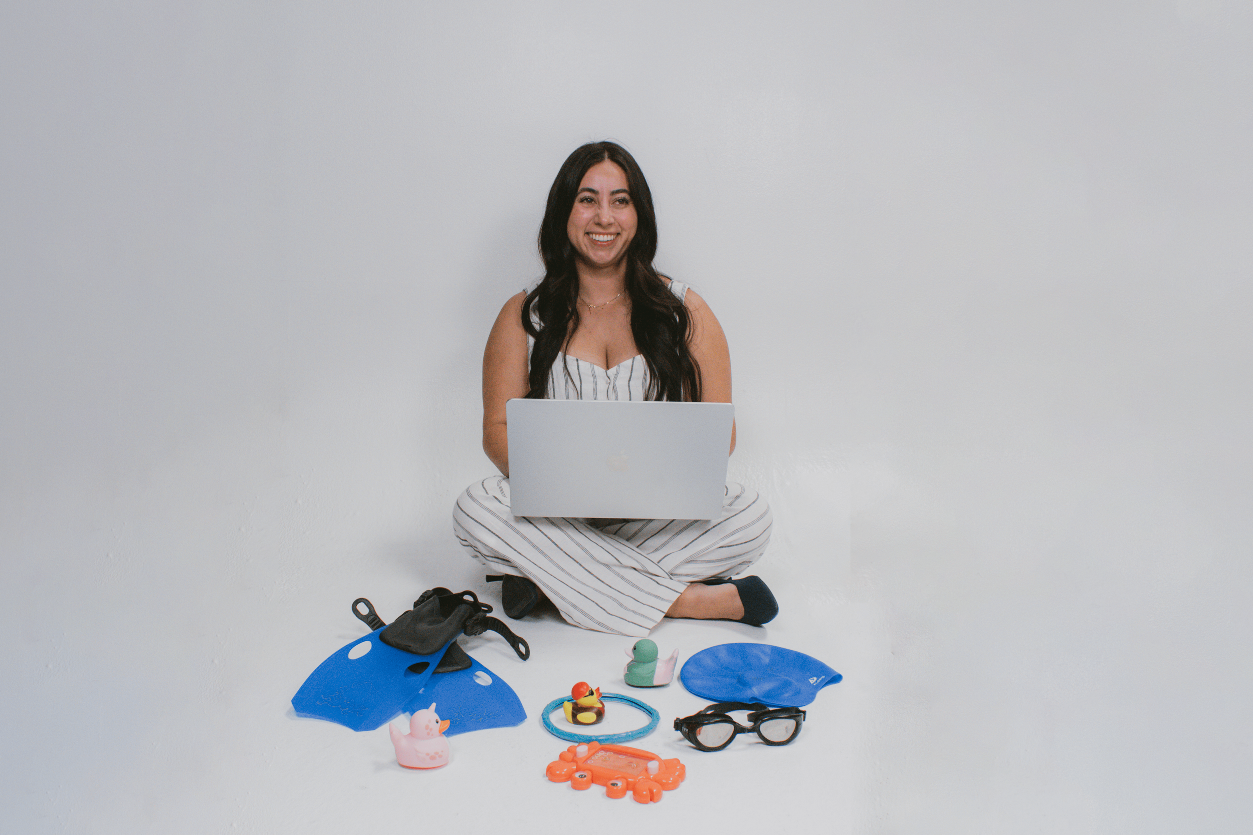 A woman sits cross-legged on the floor with a laptop on her lap, surrounded by various pool accessories and toys for Survival Swim Lessons , and smiling at the camera against a plain white wall.