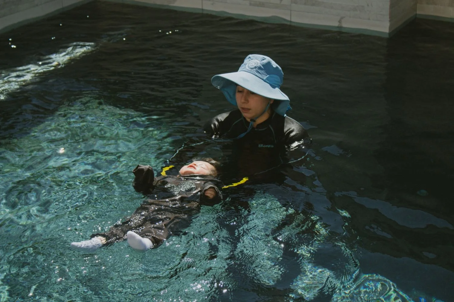 A woman wearing a blue sunhat and a black wetsuit is holding a small child in a swimming pool. The child is lying on their back with their eyes closed, wearing a black wetsuit and white socks.