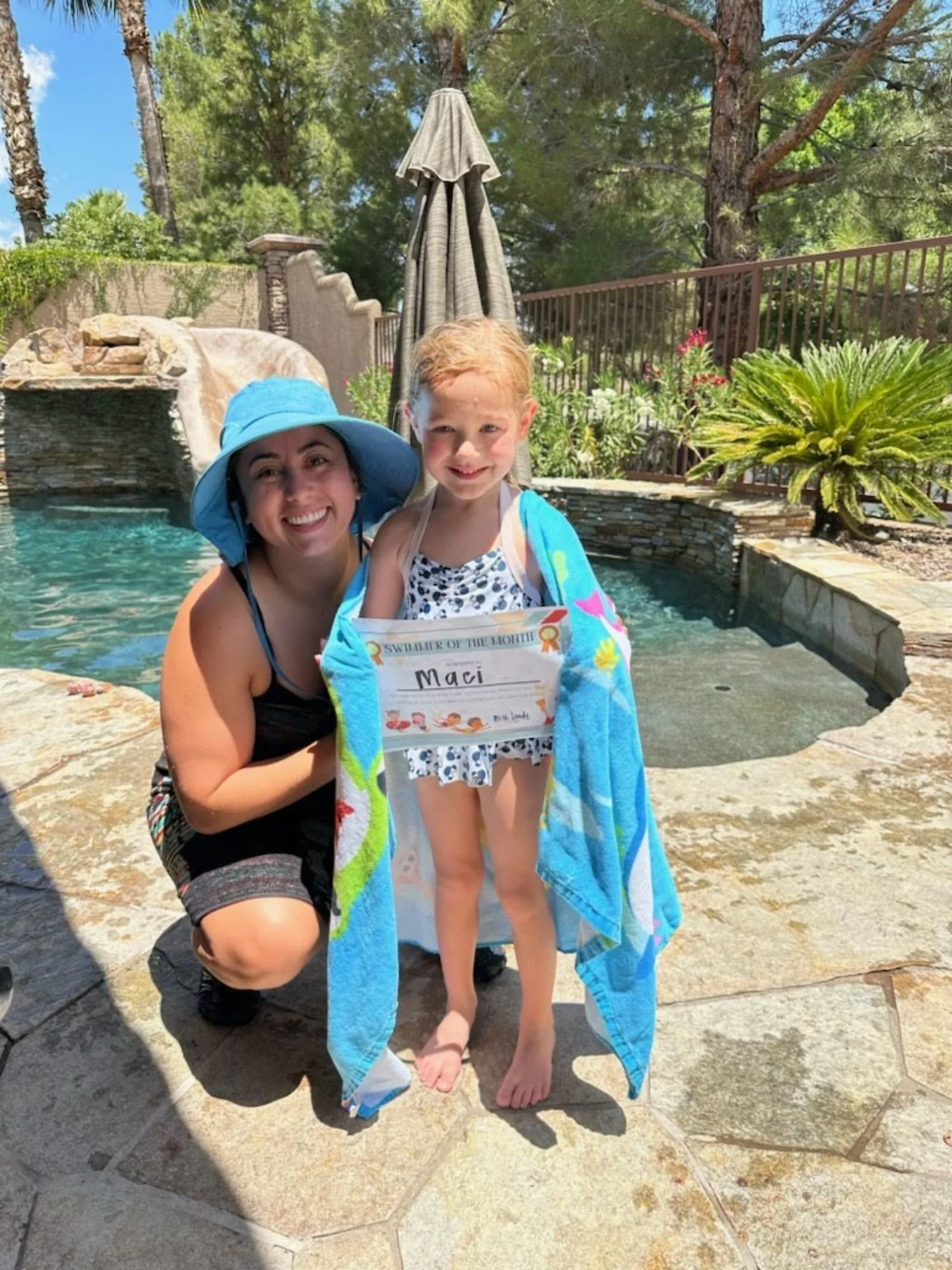 A woman and a young girl by a backyard pool. The girl is holding a sign that says 'Swimmer of the Month' with her name 'Maci' on it. The girl is wrapped in a colorful towel and is wearing a polka dot swimsuit, while the woman is wearing a black swimsuit and a blue sunhat. The background features a waterfall, trees, and a patio umbrella.