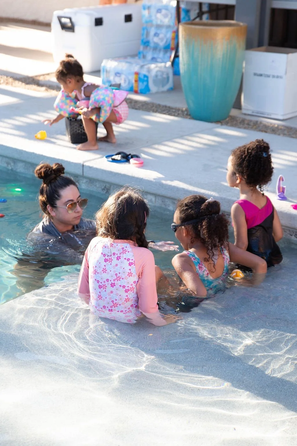 A group of women and children enjoying a swim in a backyard pool, with some sitting on the pool's edge and others in the water, while a young girl on the pool deck plays with toys nearby for Infant Survival Swim Lessons in Las Vegas
