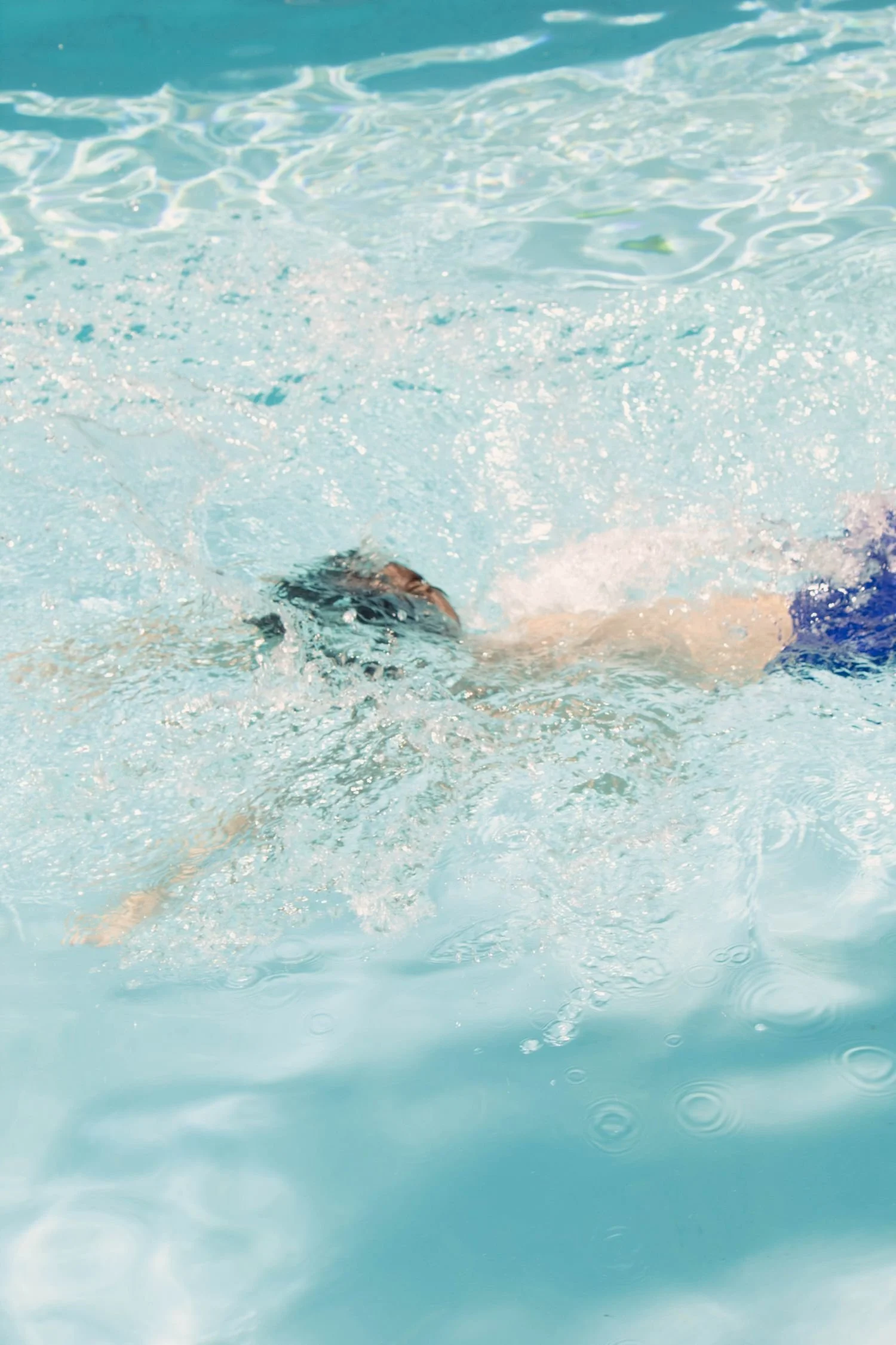 Person swimming in a pool, wearing goggles and blue swimwear, with water splashing around.  Survival Swim Lessons