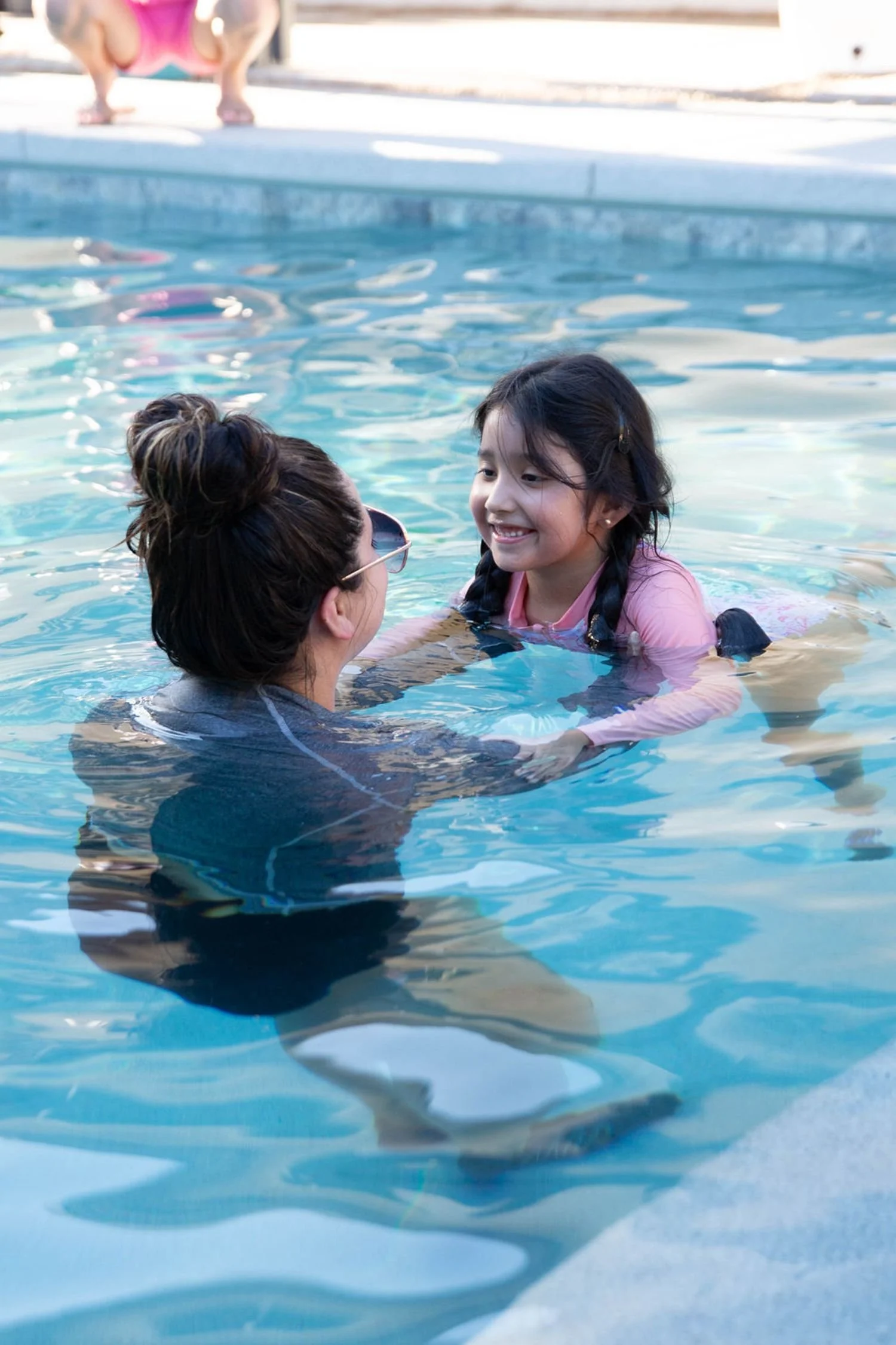 A young girl with braided hair in a pink shirt smiles as she is supported in a swimming pool by an adult woman, who is holding her hands and wearing sunglasses. Another child is visible in the background, on the edge of the pool. Survival Swim Lesson