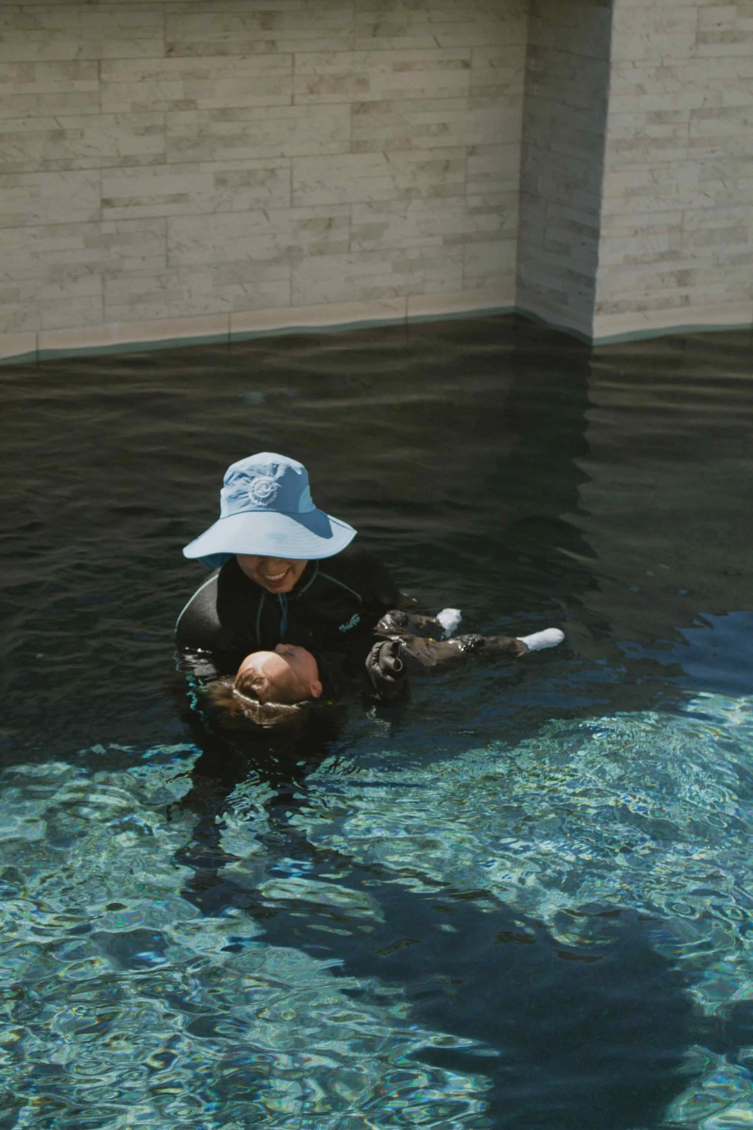 A woman in a blue sun hat and black wetsuit is joyfully holding a boy in the water, with both smiling, near a stone wall while teaching Infant Survival Swim Lessons in Las Vegas