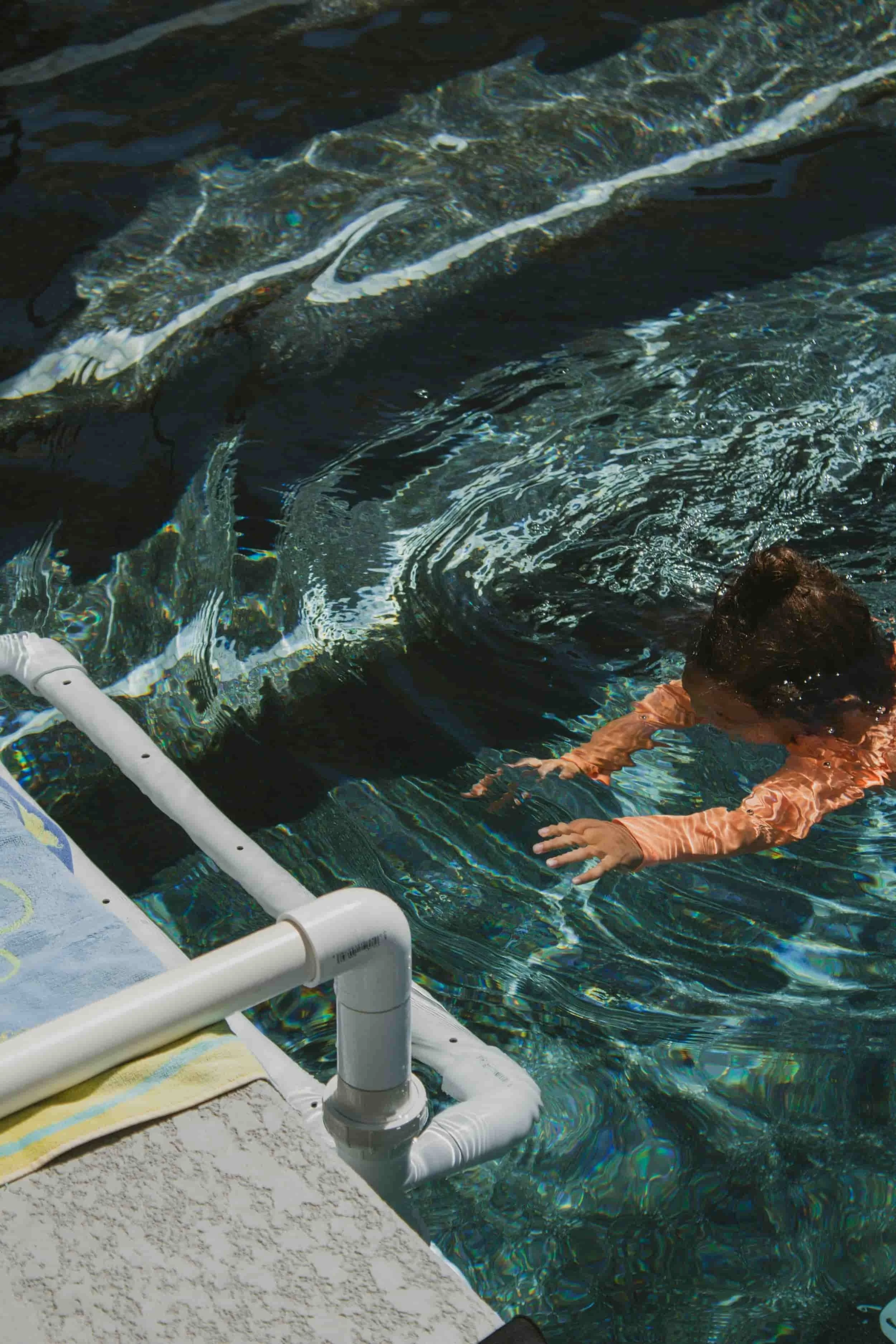 A child doing Infant Survival Swim Lessons in Las Vegas in an outdoor pool, reaching towards the water with arms extended, near a pool ladder.