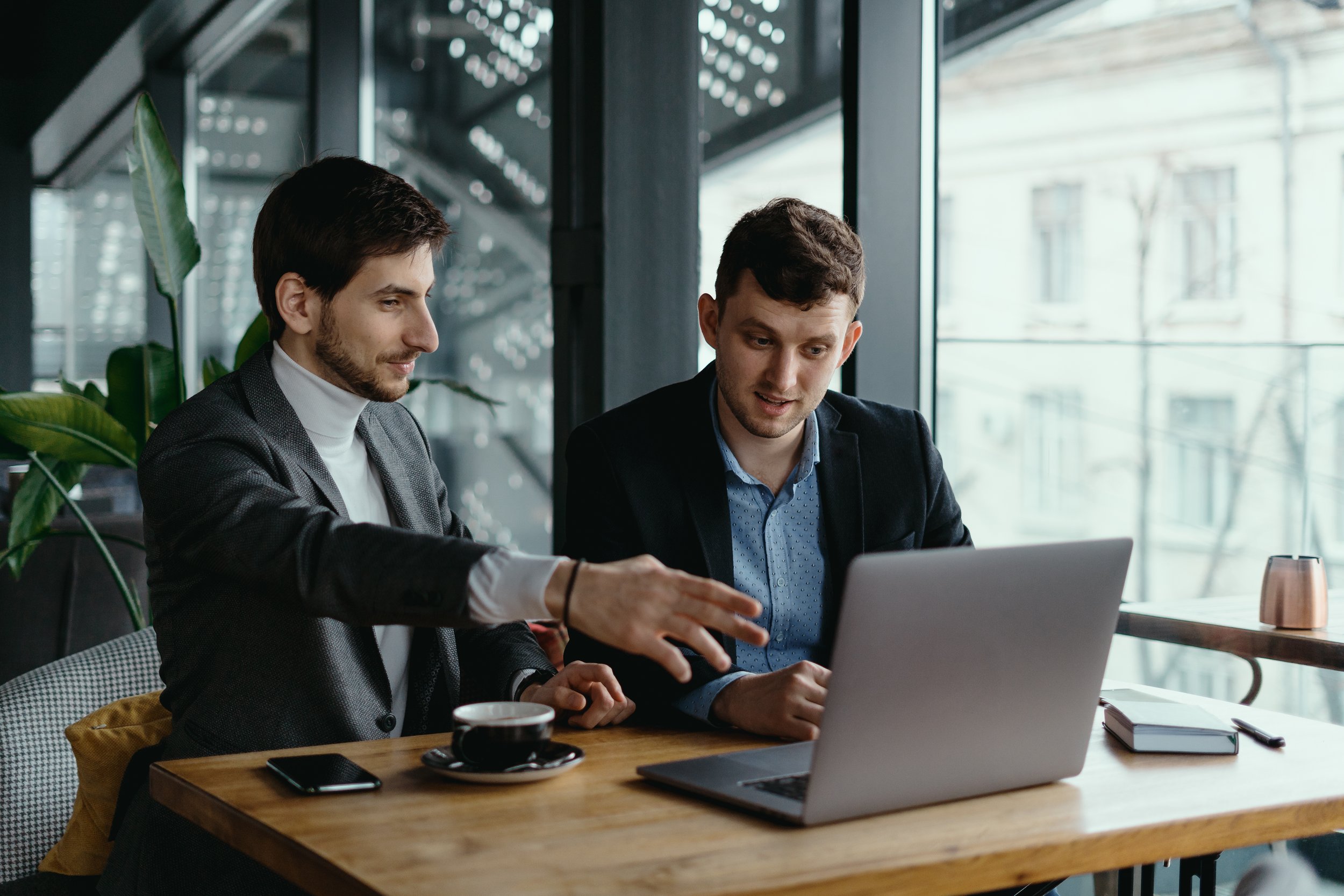 Two men in business attire working together at a wooden table with a laptop, notebook, and coffee in a modern office with large windows and natural light.