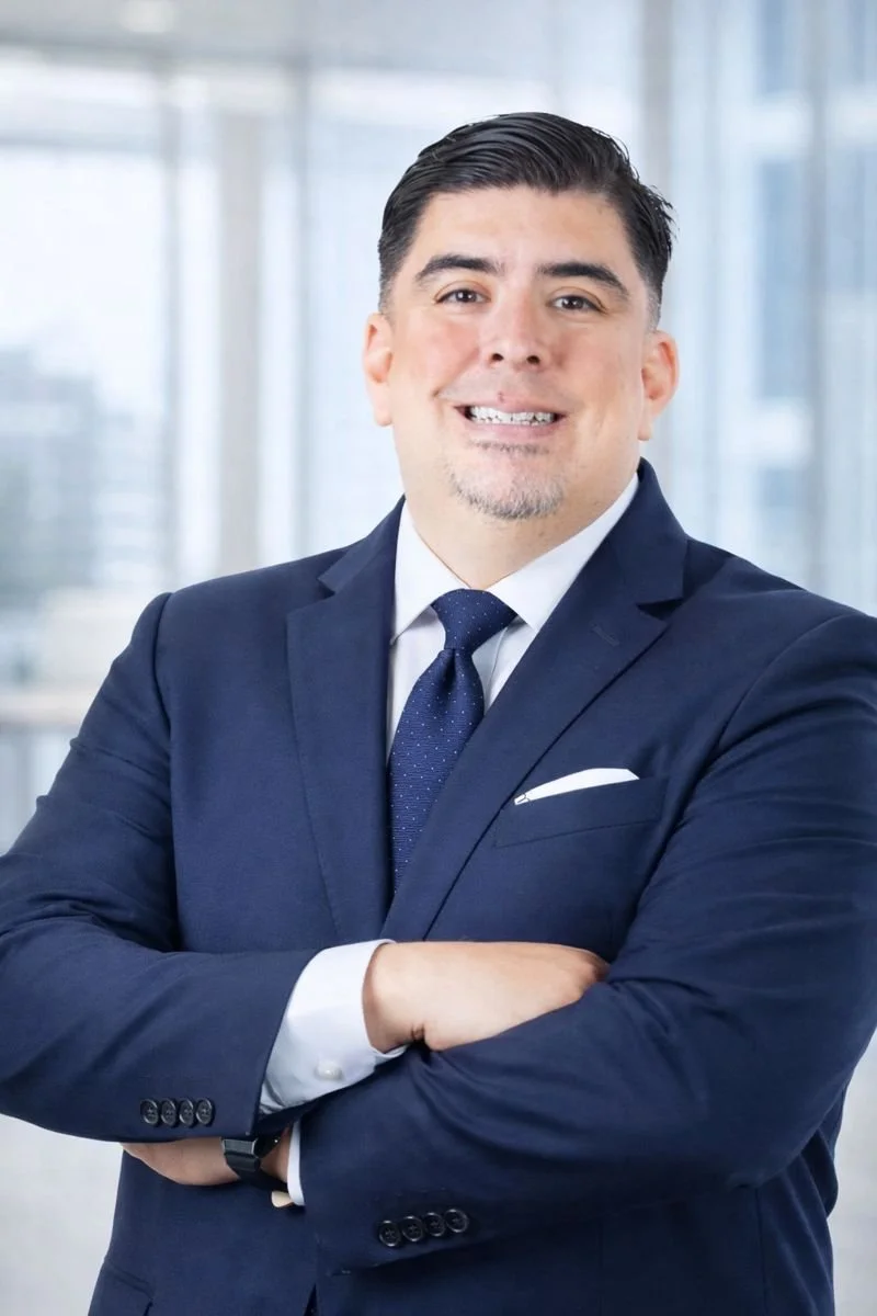 Portrait of a professional man wearing a navy blue suit, white shirt, and blue tie, standing with arms crossed in a modern office setting with large windows.