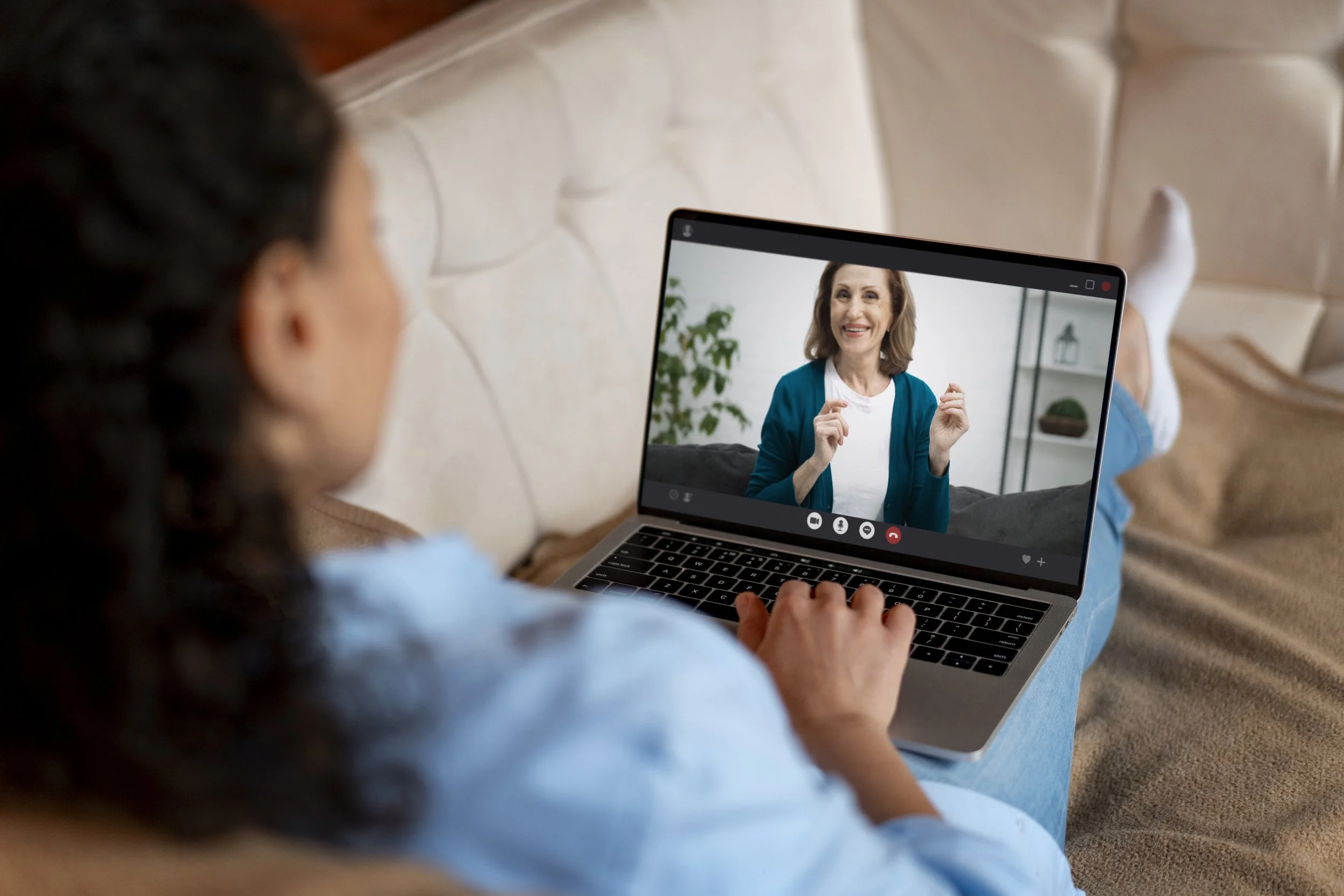 A woman on a laptop screen during a video call with an older woman smiling and making gestures. The woman on the laptop is blurred but the woman on the screen is clear, wearing a blue cardigan and white shirt, sitting in a cozy room with a plant and decorative items in the background.