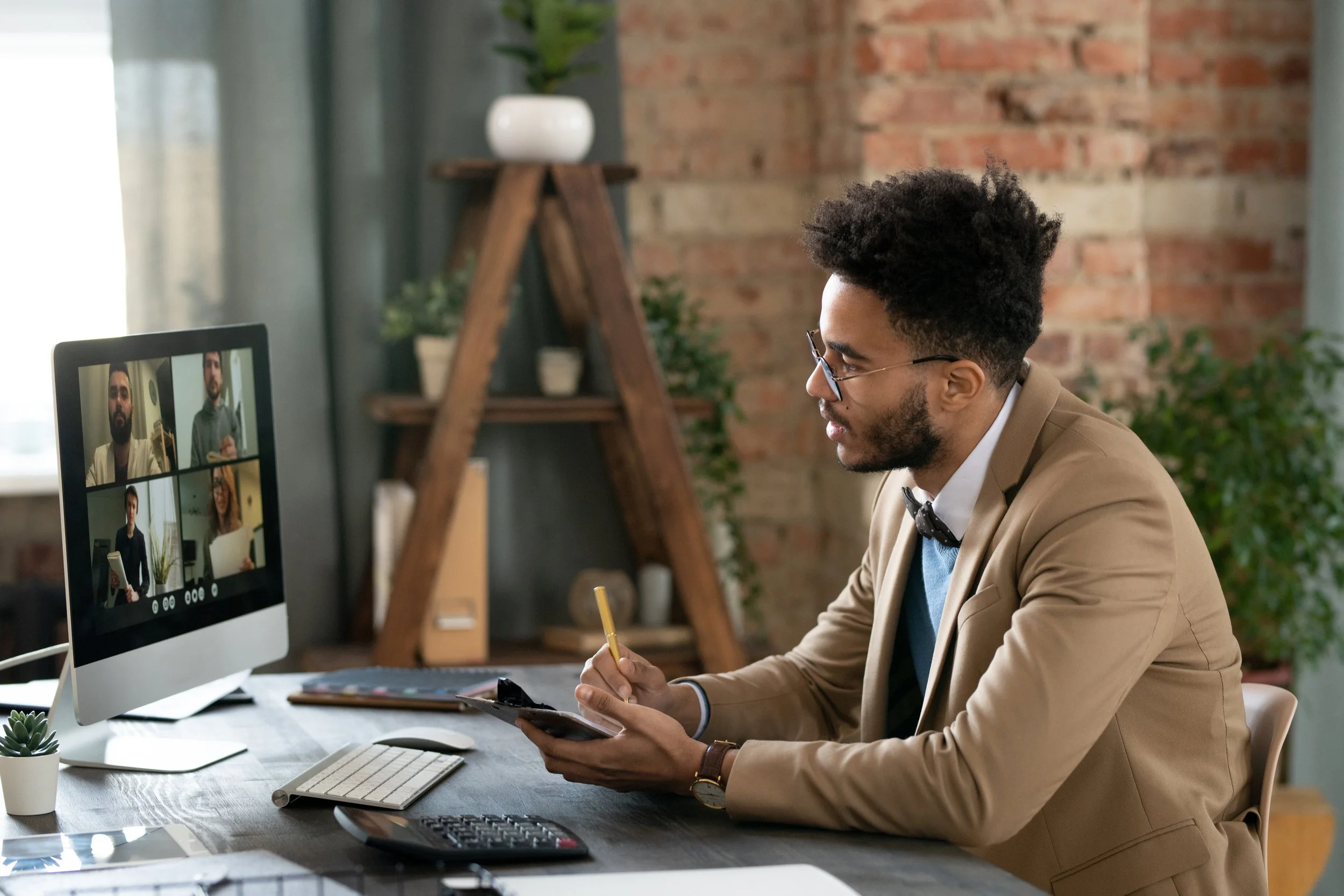 A man in a beige blazer and glasses is sitting at a desk, participating in a video conference on a desktop computer, with multiple people visible on the screen.