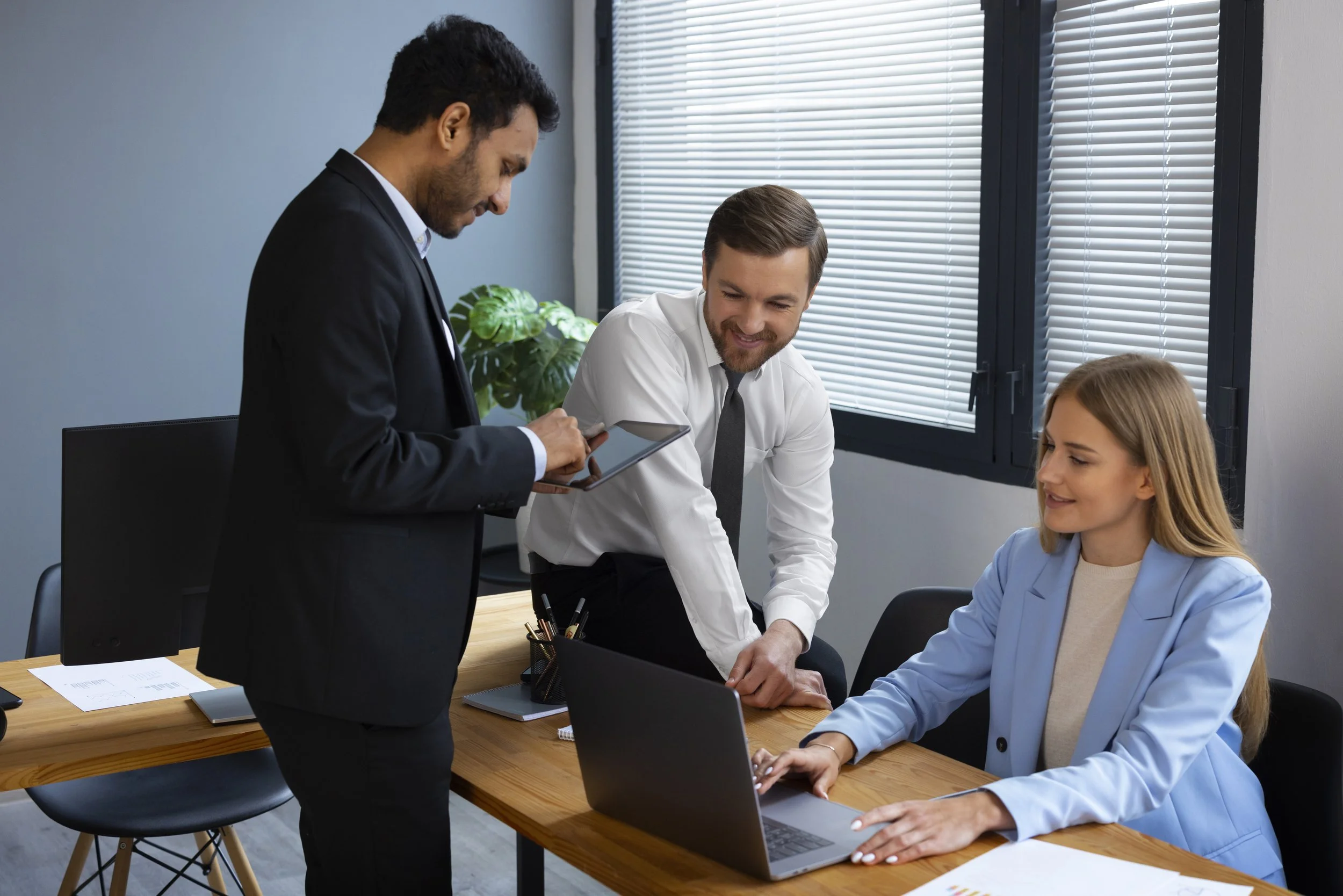 Three professionals in business attire sharing ideas and collaborating around a conference table in an office with large windows and blinds.