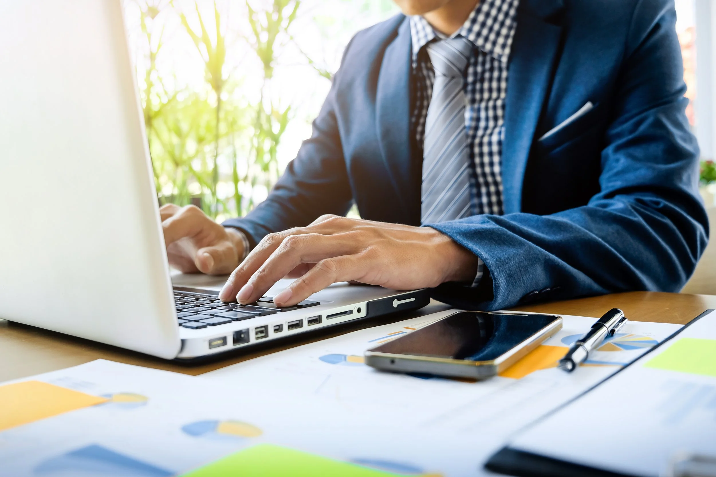 A businessman in a dark blue suit, checkered shirt, and striped tie working on a silver laptop at a desk with papers, a smartphone, and a pen, with a background of greenery and sunlight.
