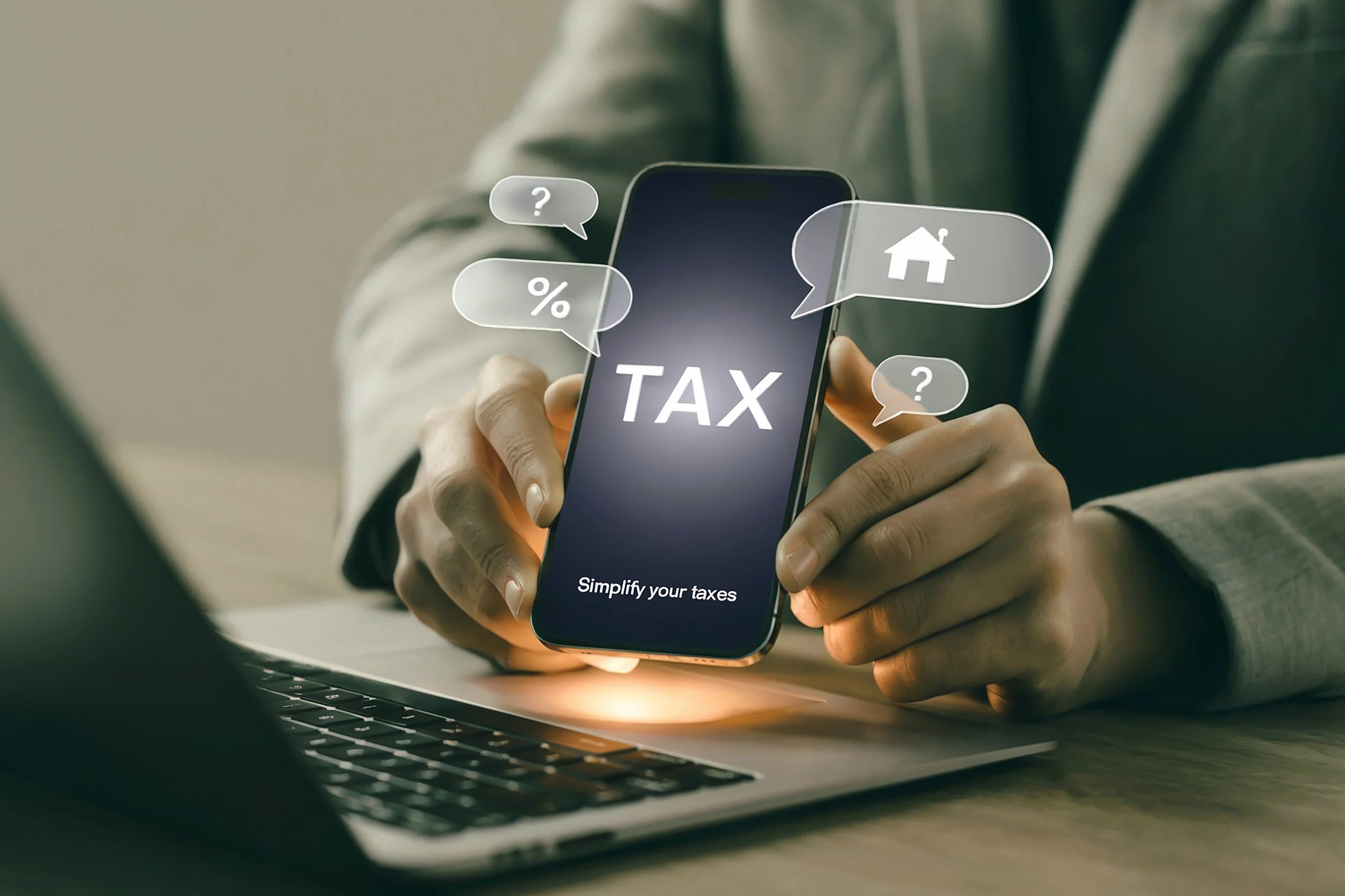 Person holding a smartphone with the word 'TAX' on the screen and speech bubbles with question marks, a house, and a percentage symbol, while a laptop is on the table.