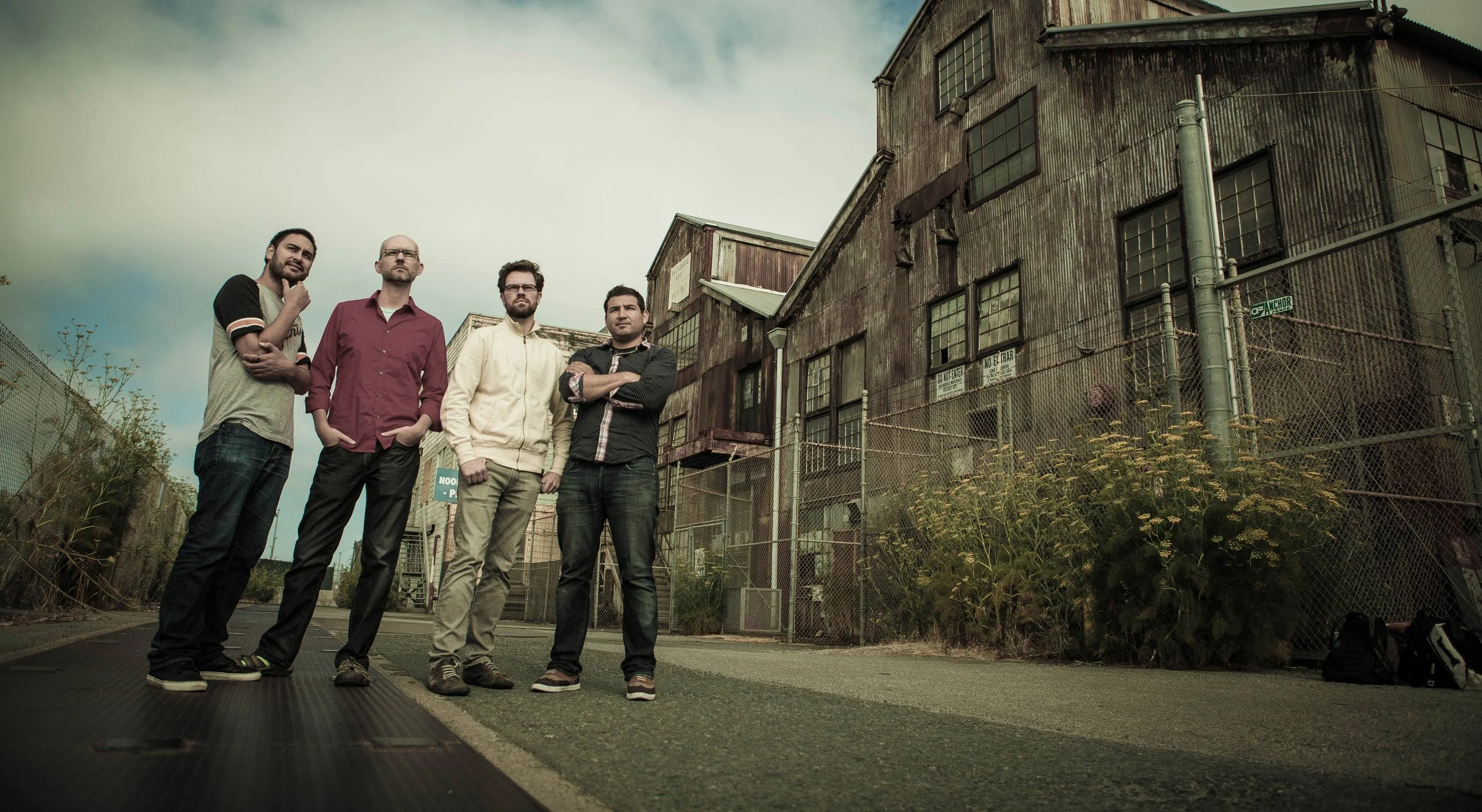 Four men standing on a street in front of an old, weathered industrial building with overgrown plants, under a cloudy sky.