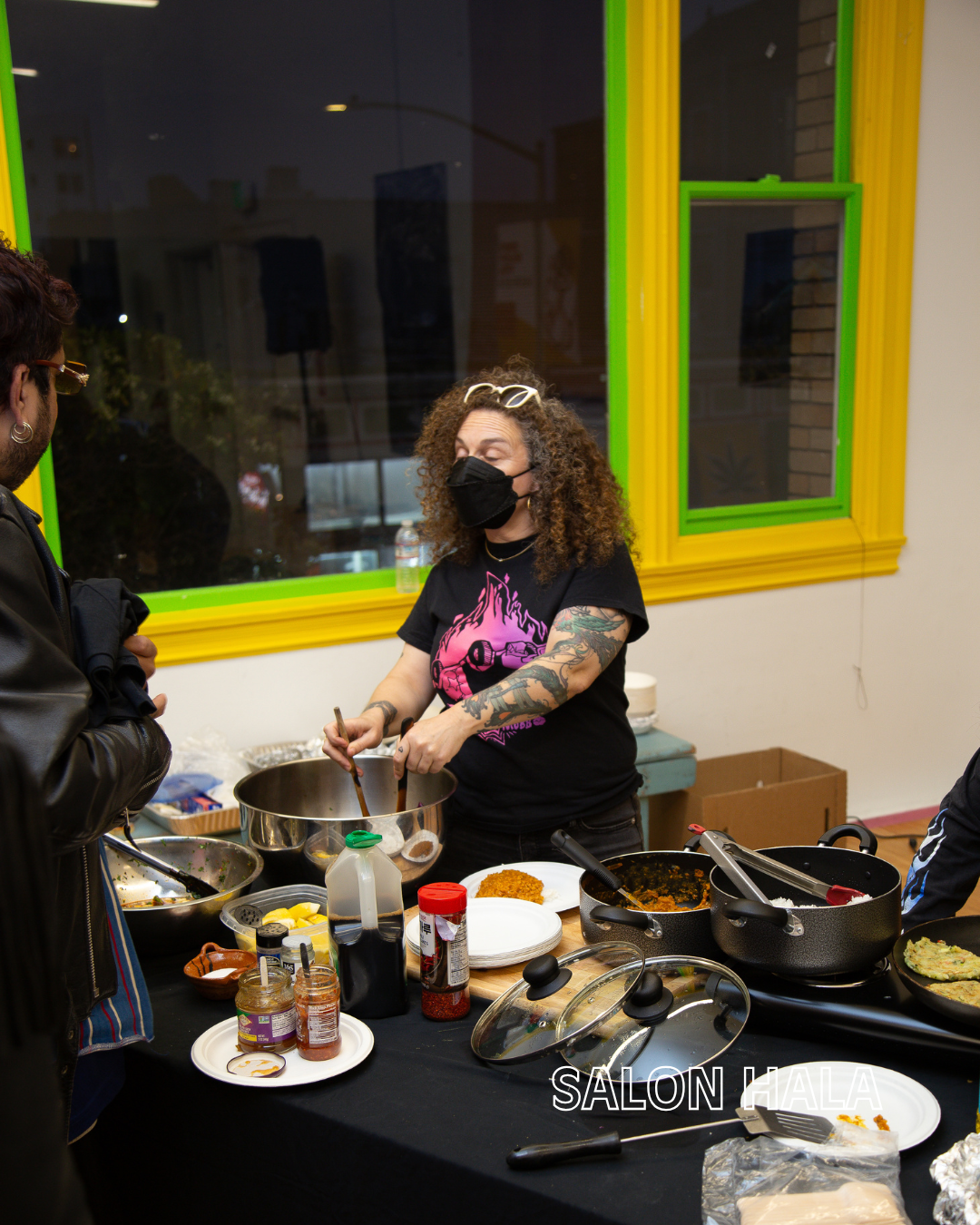 A woman with curly hair, black mask, and sunglasses on her head cooking at a food stand or table. She is stirring a pot with various ingredients around, while a person in a black jacket listens. The background has a window with green and yellow borde