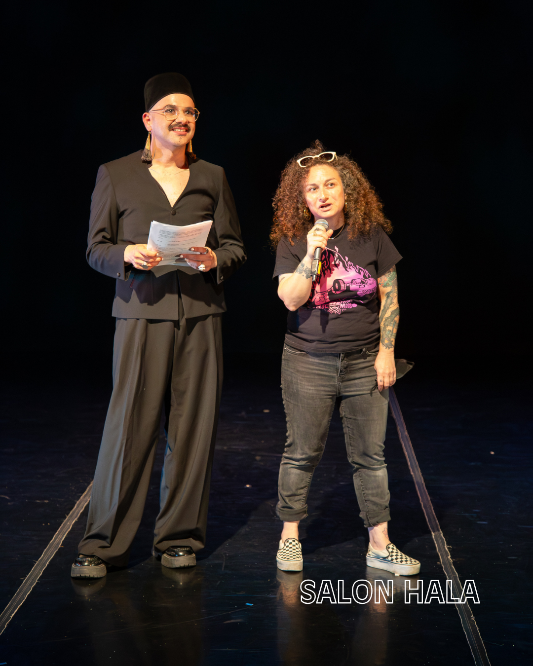 Two women speaking on stage at SALON HALA event, with one holding a microphone and the other holding papers, against a dark background.