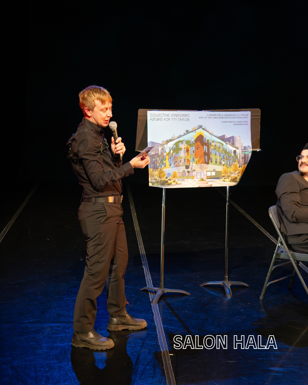 A person wearing black protective gear and boots is standing on stage holding a microphone and reading from a paper, next to a presentation board displaying an architectural rendering of a building with the title "Collective Envisioning: Futures for 
