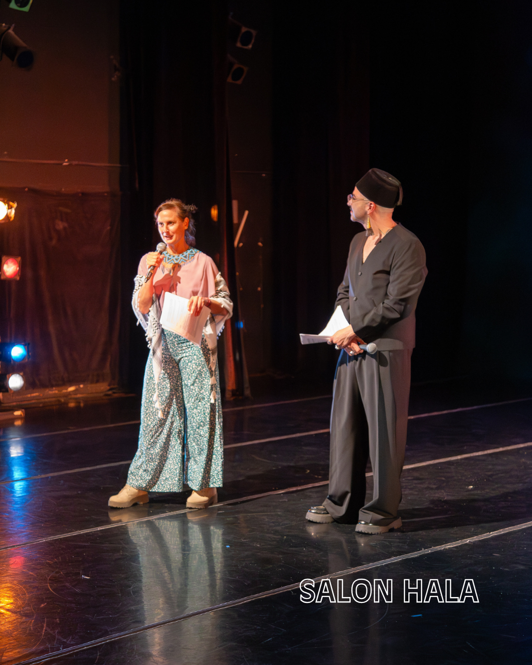 Two people on stage, one woman speaking into a microphone and holding papers, the other man wearing traditional Muslim attire with a cap, both holding papers, stage with black curtains and colorful lights, labeled 'SALON HALA'.