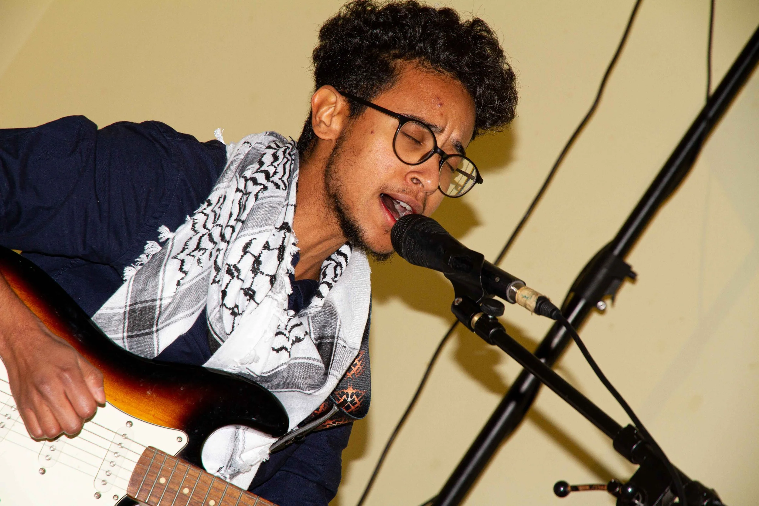 Man with curly black hair, glasses, and a beard singing into a microphone while playing an electric guitar in front of a yellow wall.