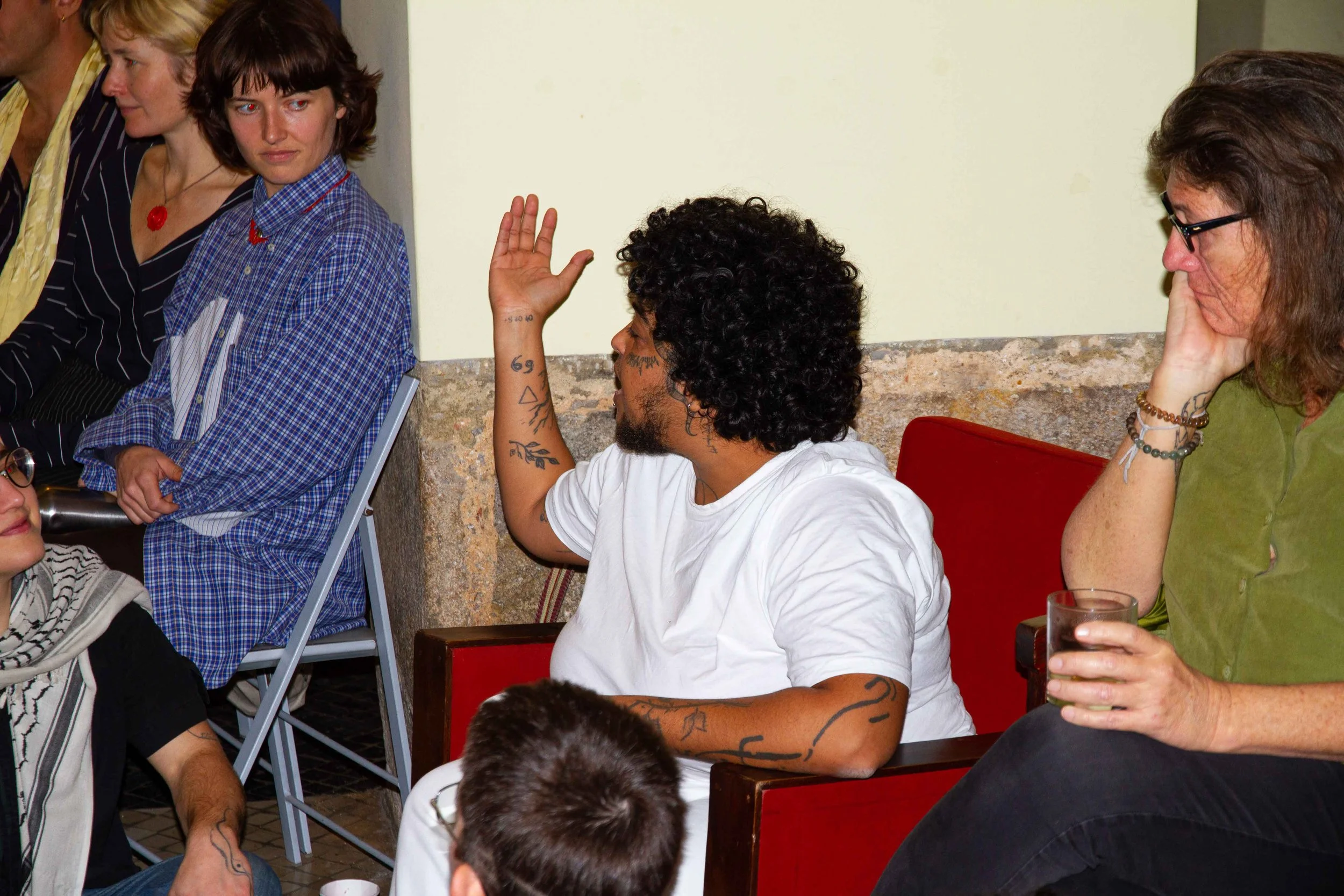 A group of people sitting around a table in a room, engaging in a discussion. One person with curly hair and tattoos is raising their hand, while others listen attentively. The scene appears to be an informal meeting or discussion.