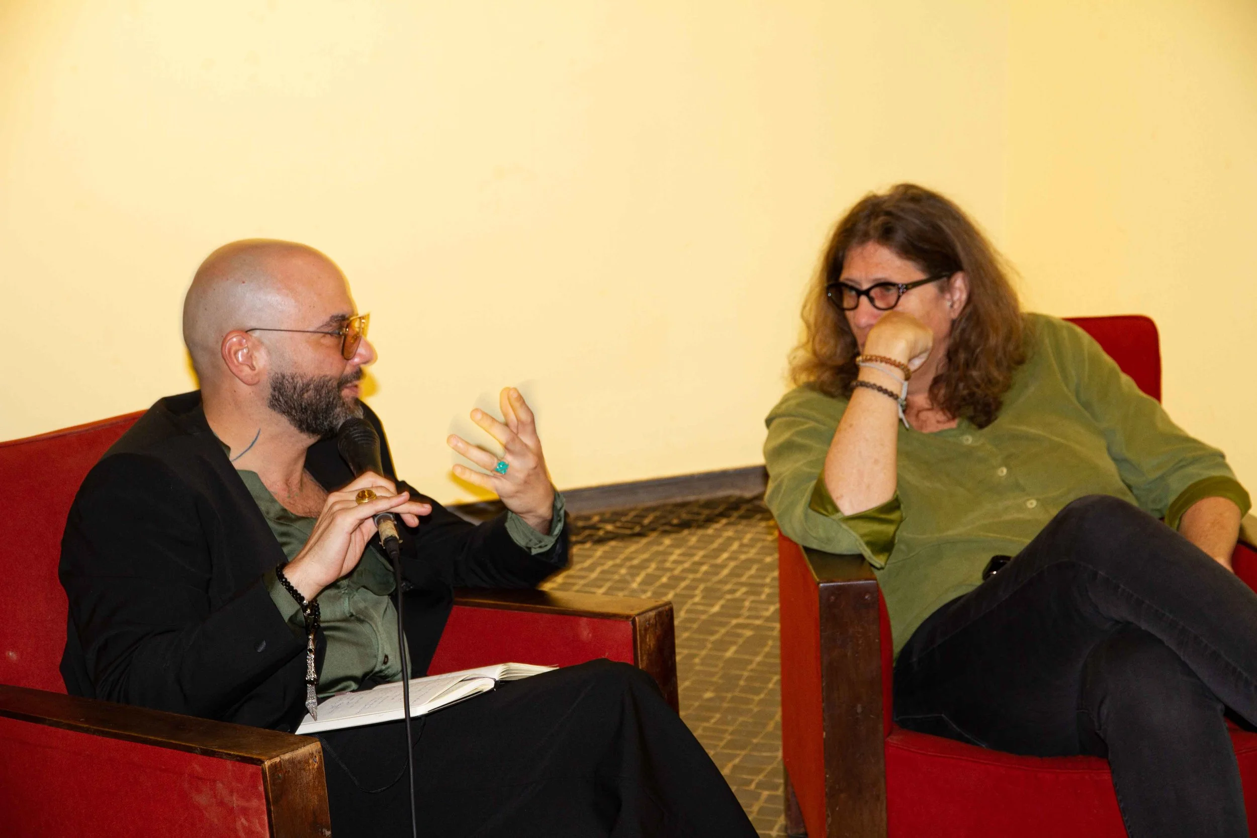 Two people sitting and talking in a room with yellow walls. The person on the left has a beard, glasses, and a shaved head, and is holding a microphone. The person on the right has long brown hair, glasses, and is wearing a green shirt, resting her c