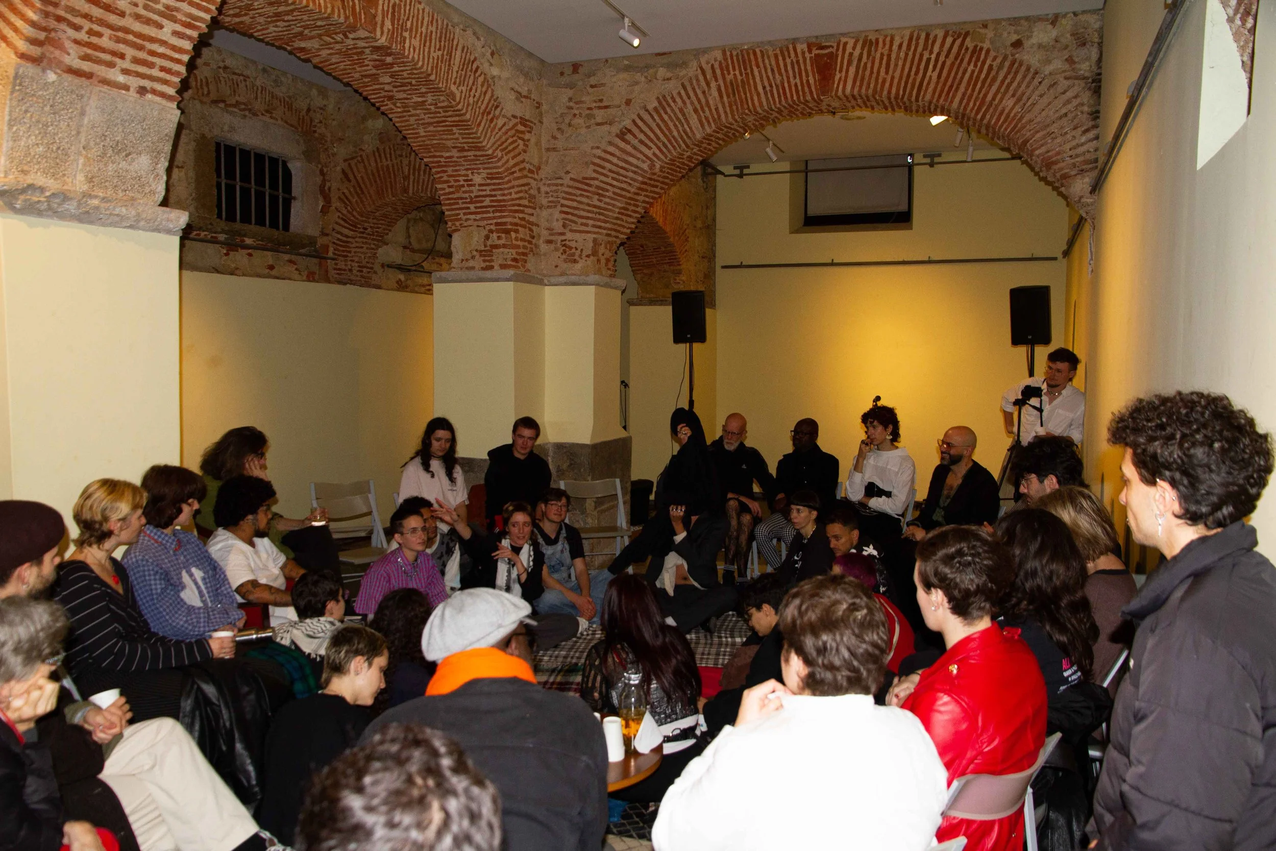 A large group of people gathered in a room with exposed brick arches and yellow walls, sitting and standing, engaged in an event or discussion.