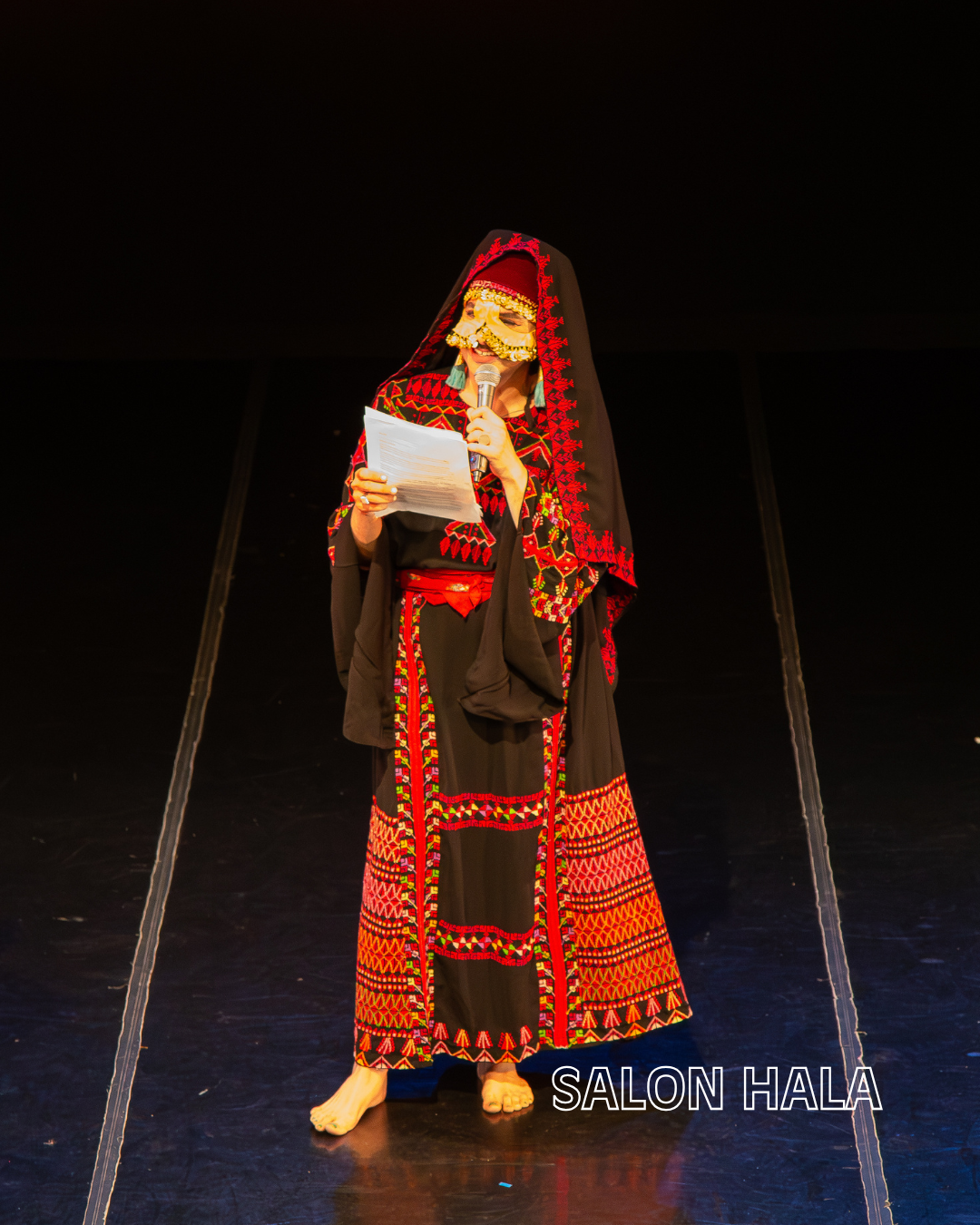 Person wearing traditional indigenous attire and mask, holding a microphone and reading from papers on stage at SALON HALA event.