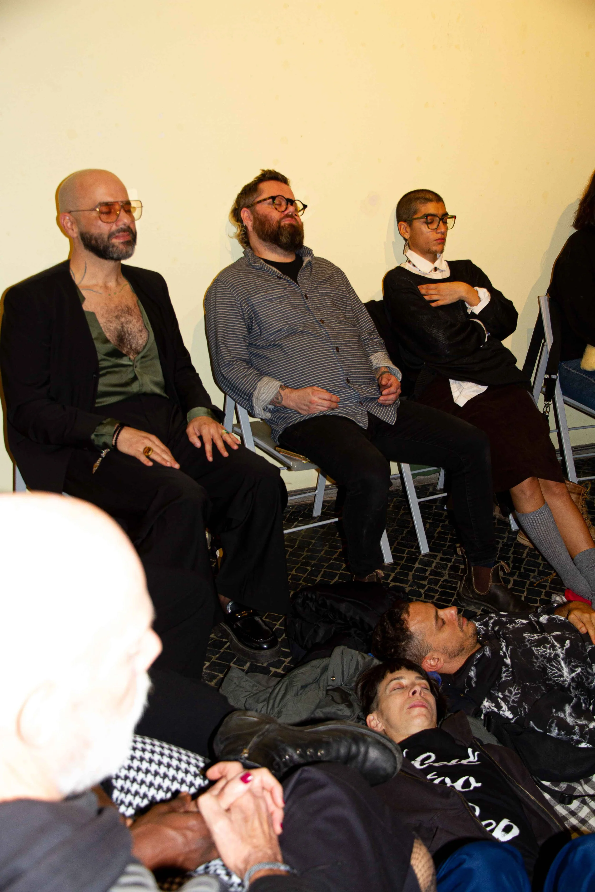 Group of people sitting and lying on the floor with eyes closed, possibly meditating or resting at an indoor event.