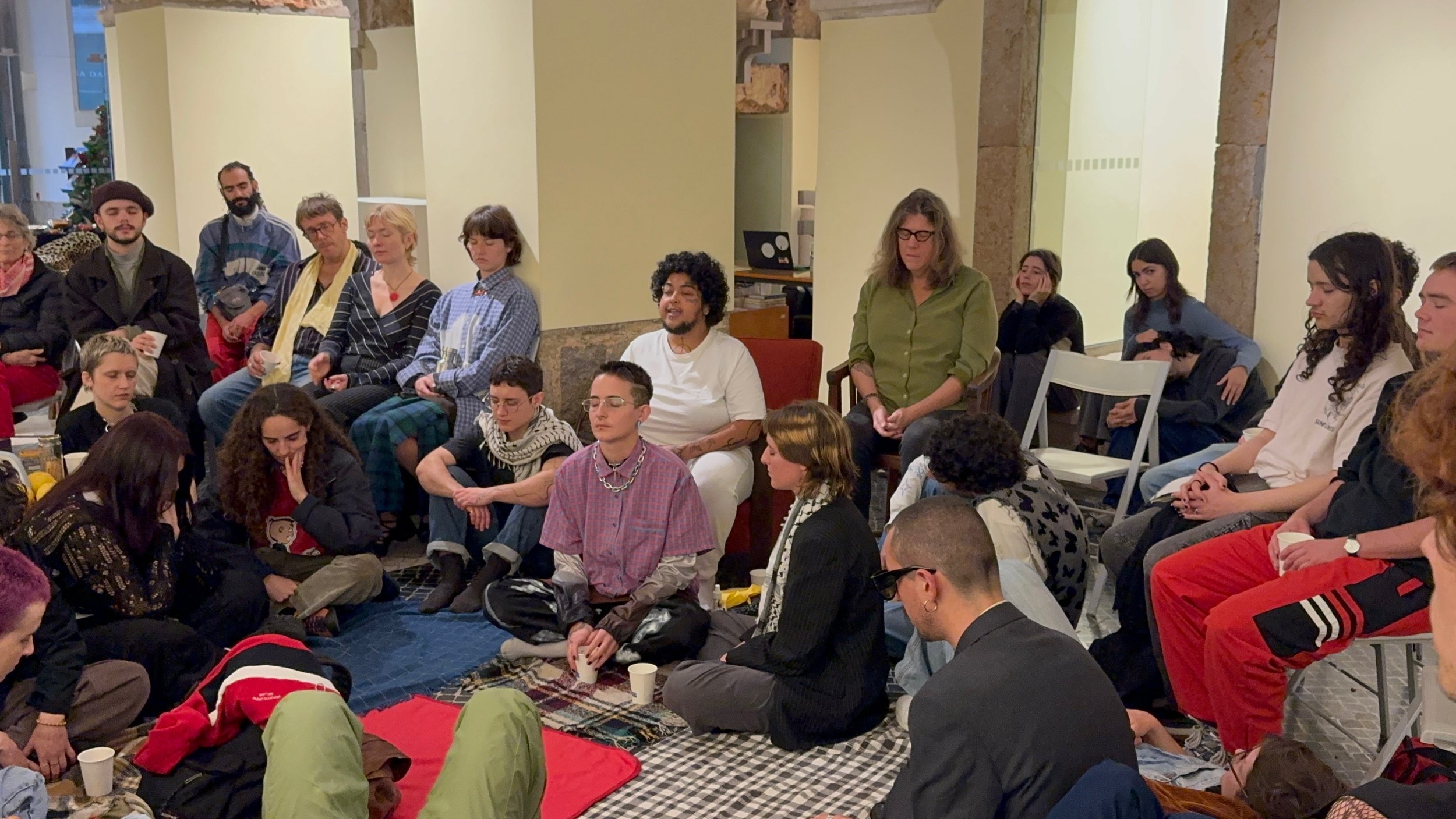 A diverse group of people sitting and standing in a room, engaging in a meditation or mindfulness session. Some have their eyes closed, others are looking around or at the speaker. The setting appears to be indoors, with a decorated Christmas tree vi