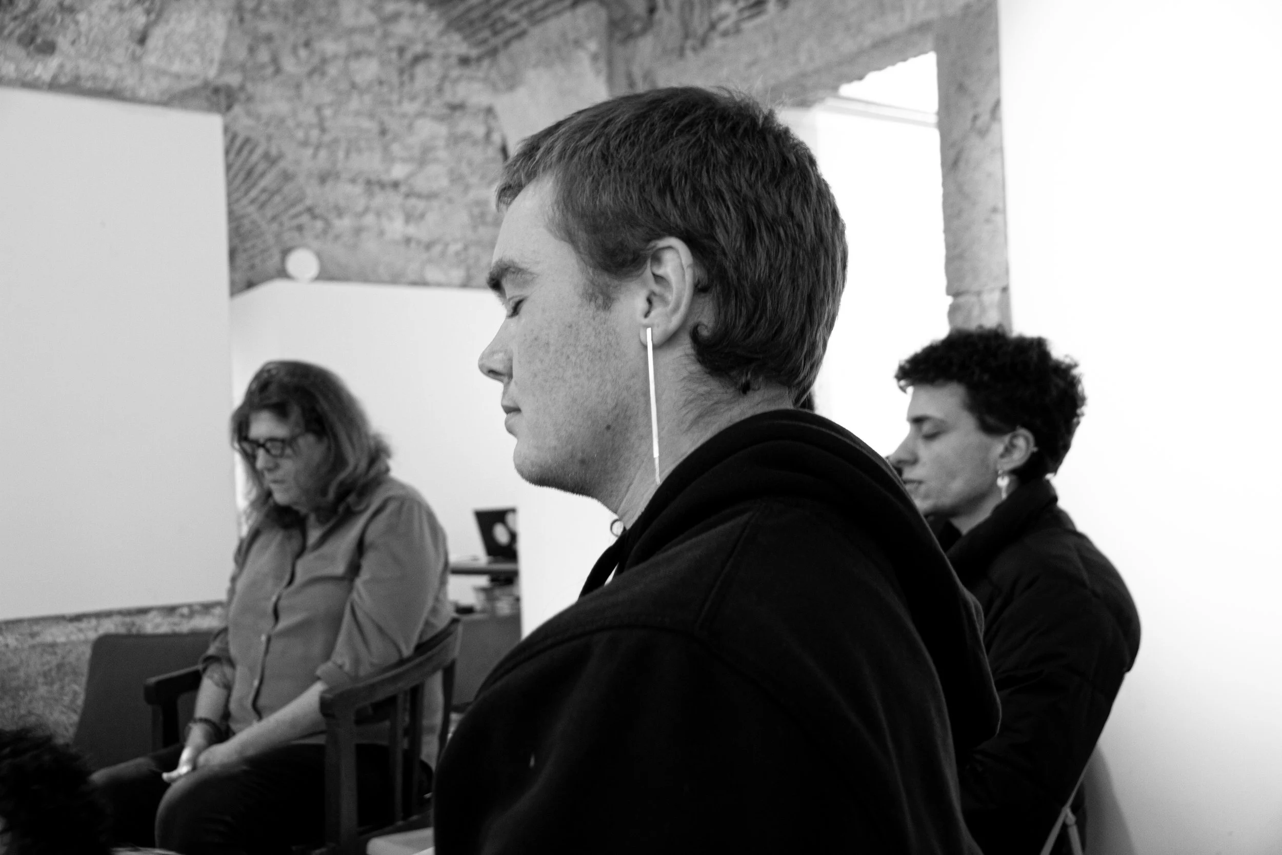 Three people sitting in a row with their eyes closed, possibly meditating or praying, in a room with exposed brick walls.