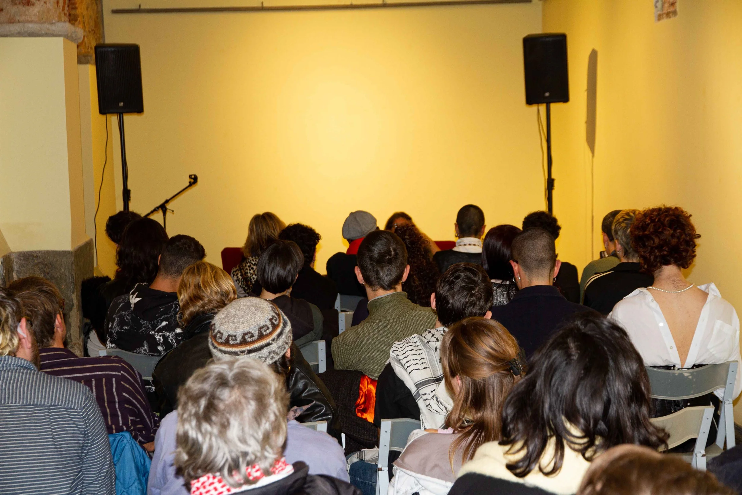 Audience gathered in a room with a yellow wall, listening to a speaker, with two black speakers mounted high on the wall and a microphone stand visible.