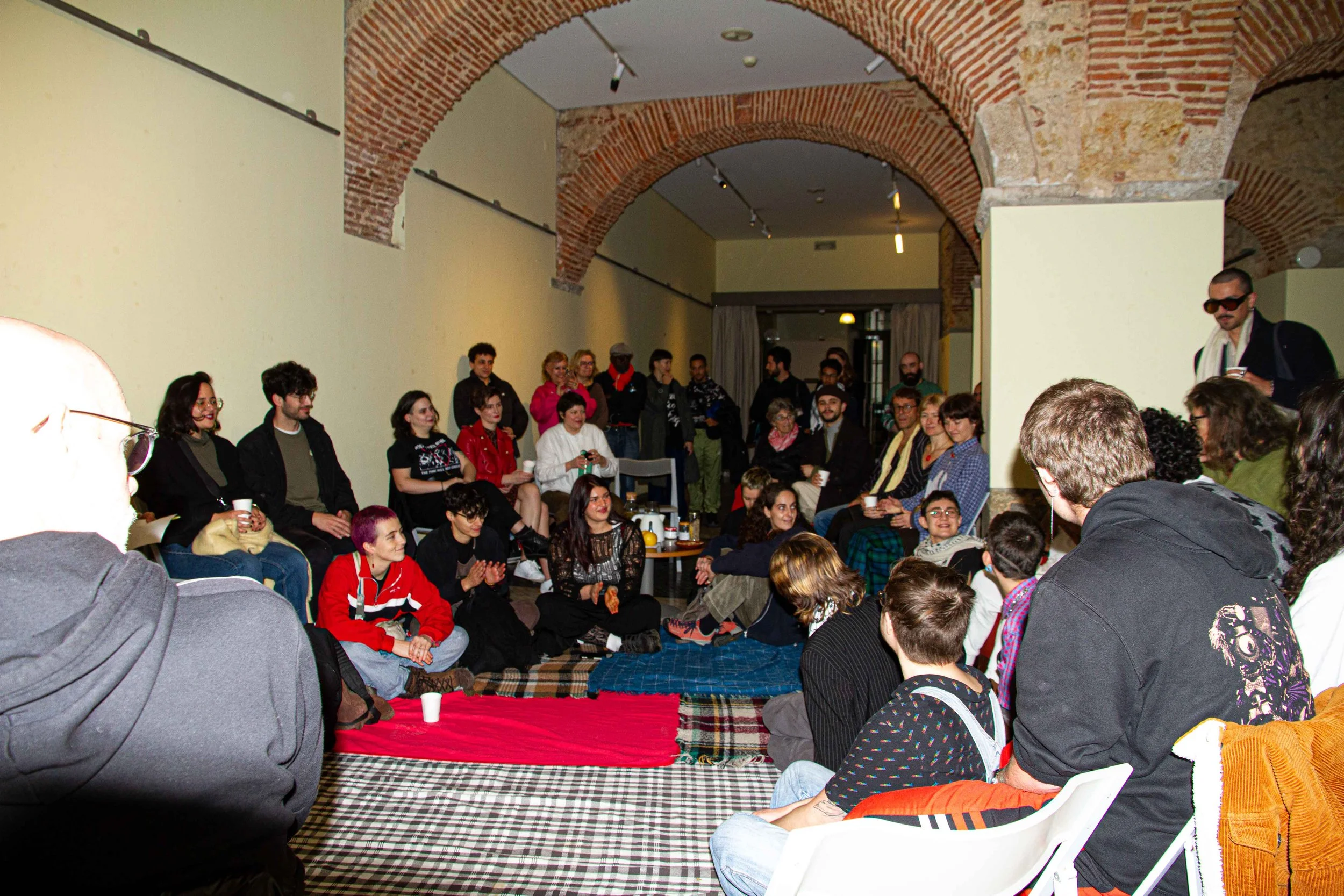 A diverse group of people gathered indoors under brick arches, some sitting on the floor, others standing, engaged in an event or discussion, with a predominantly warm-toned interior.