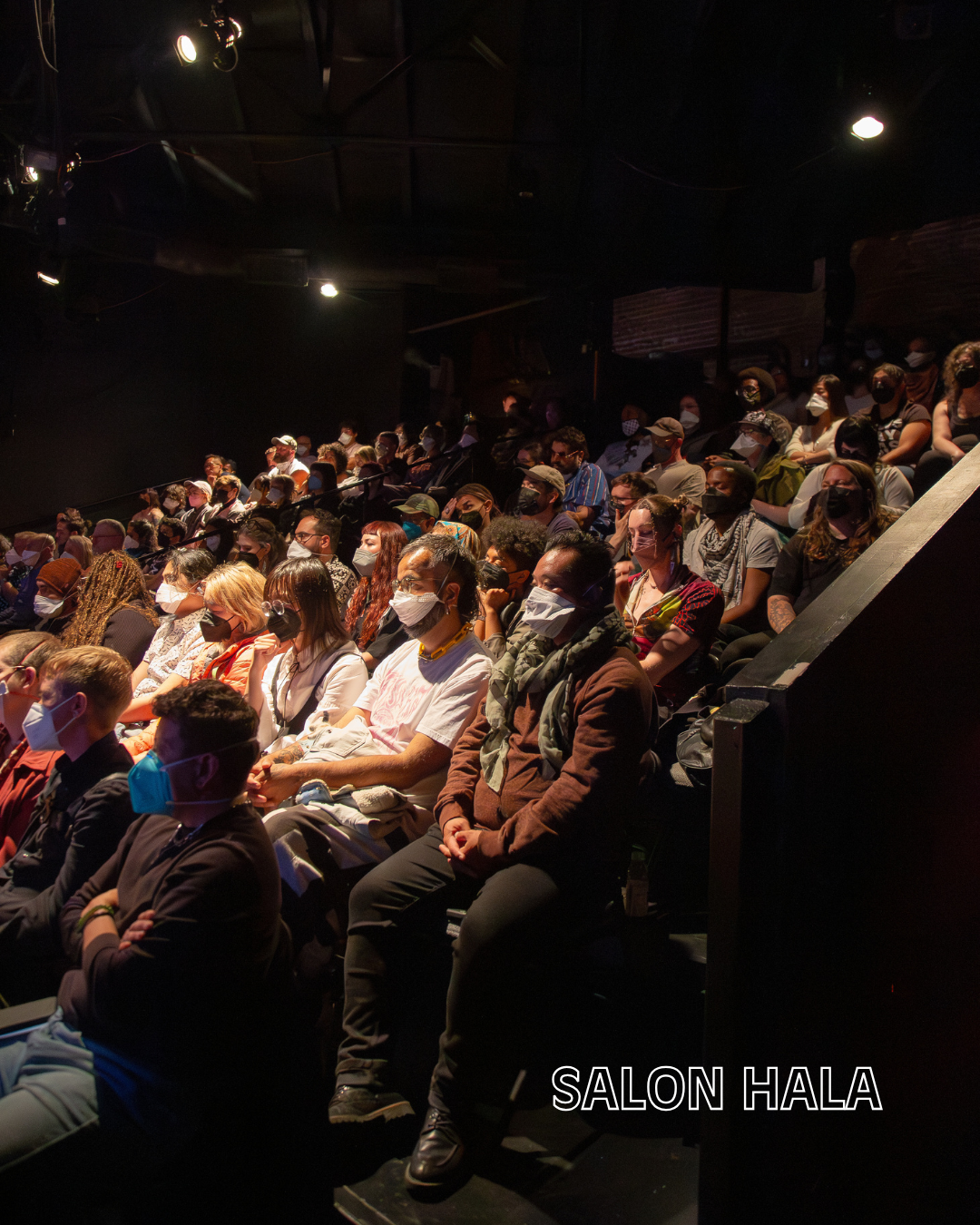 An audience wearing face masks seated in a dark theater at a performance or presentation. The text 'SALON HALA' is visible at the bottom right of the image.
